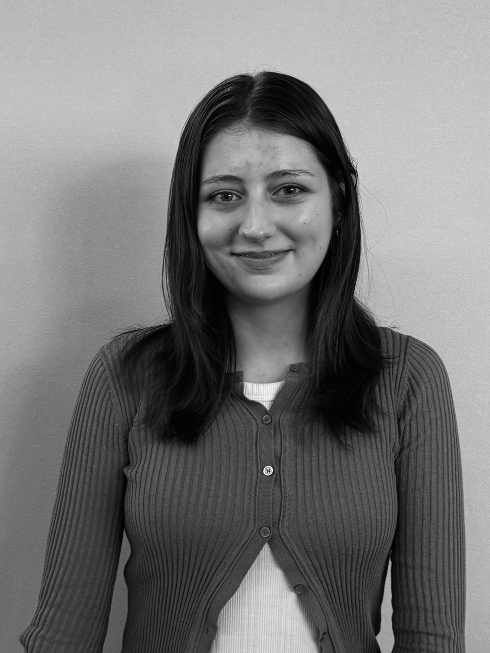 Black and white portrait of a young woman with wavy dark hair wearing a light-colored off-shoulder blouse, smiling gently, standing against a plain background.