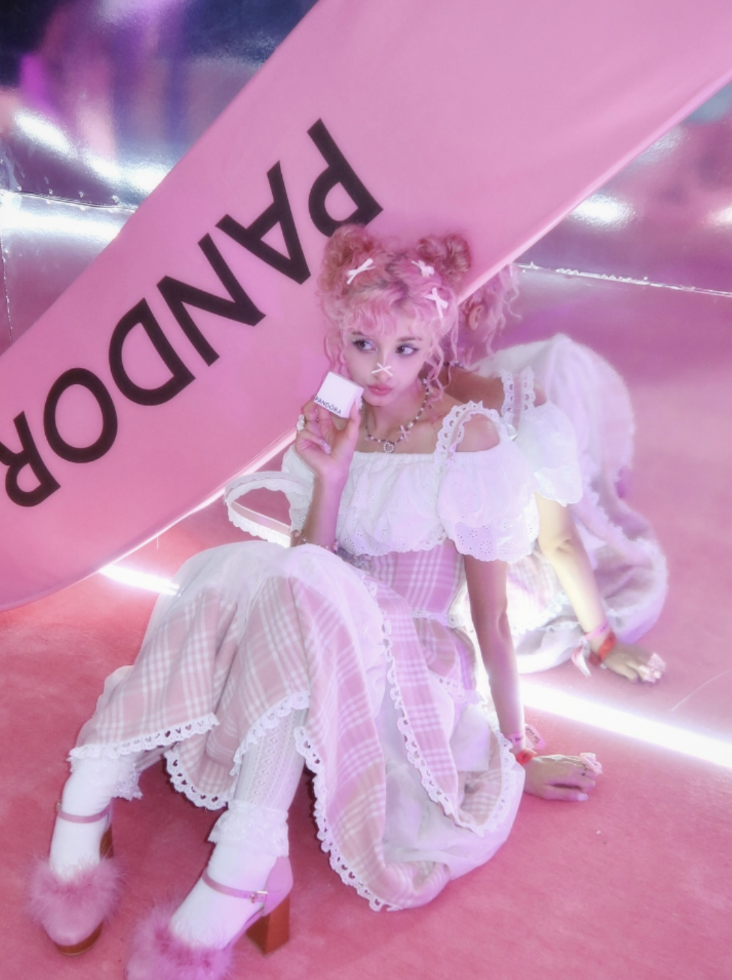 Young woman with pink curly hair styled in two buns, wearing a white floral dress with pink plaid accents, white lace socks, and fluffy pink shoes. She is holding a small box labeled 'Pandora' and sitting on a pink carpeted floor in front of a pink and metallic background, with the word 'PANDORA' visible upside down.