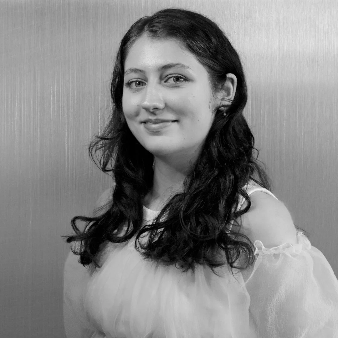 Black and white portrait of a young woman with wavy dark hair wearing a light-colored off-shoulder blouse, smiling gently, standing against a plain background.