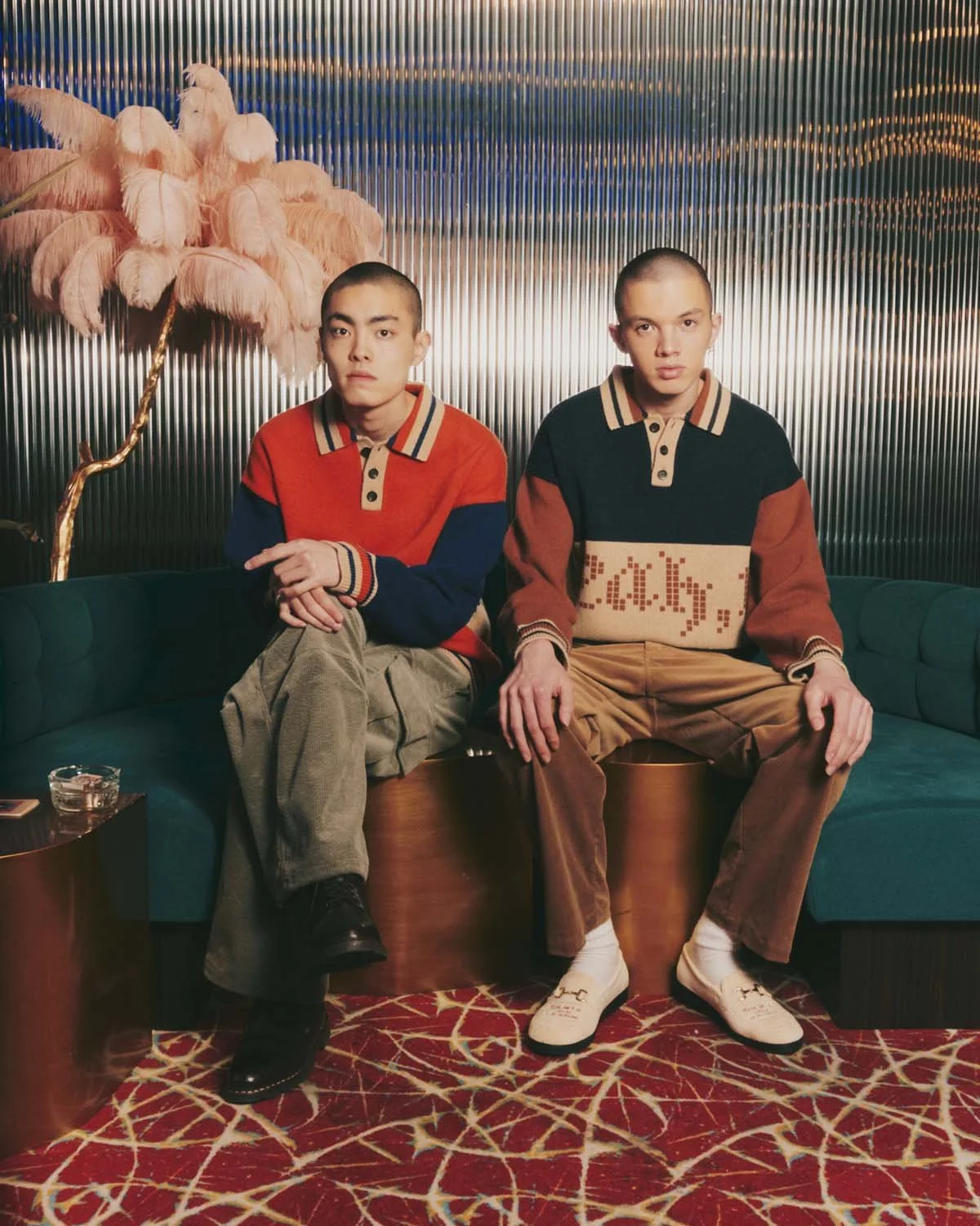 Two young men sitting on a teal couch in a stylish room with a metallic wall and a pink feathered lamp in the background. One wears a red, blue, and beige sweater, khaki pants, and black boots. The other wears a black, beige, and rust-colored sweater, brown pants, and white shoes. There is a small black table with a glass of water beside them and a red patterned carpet on the floor.