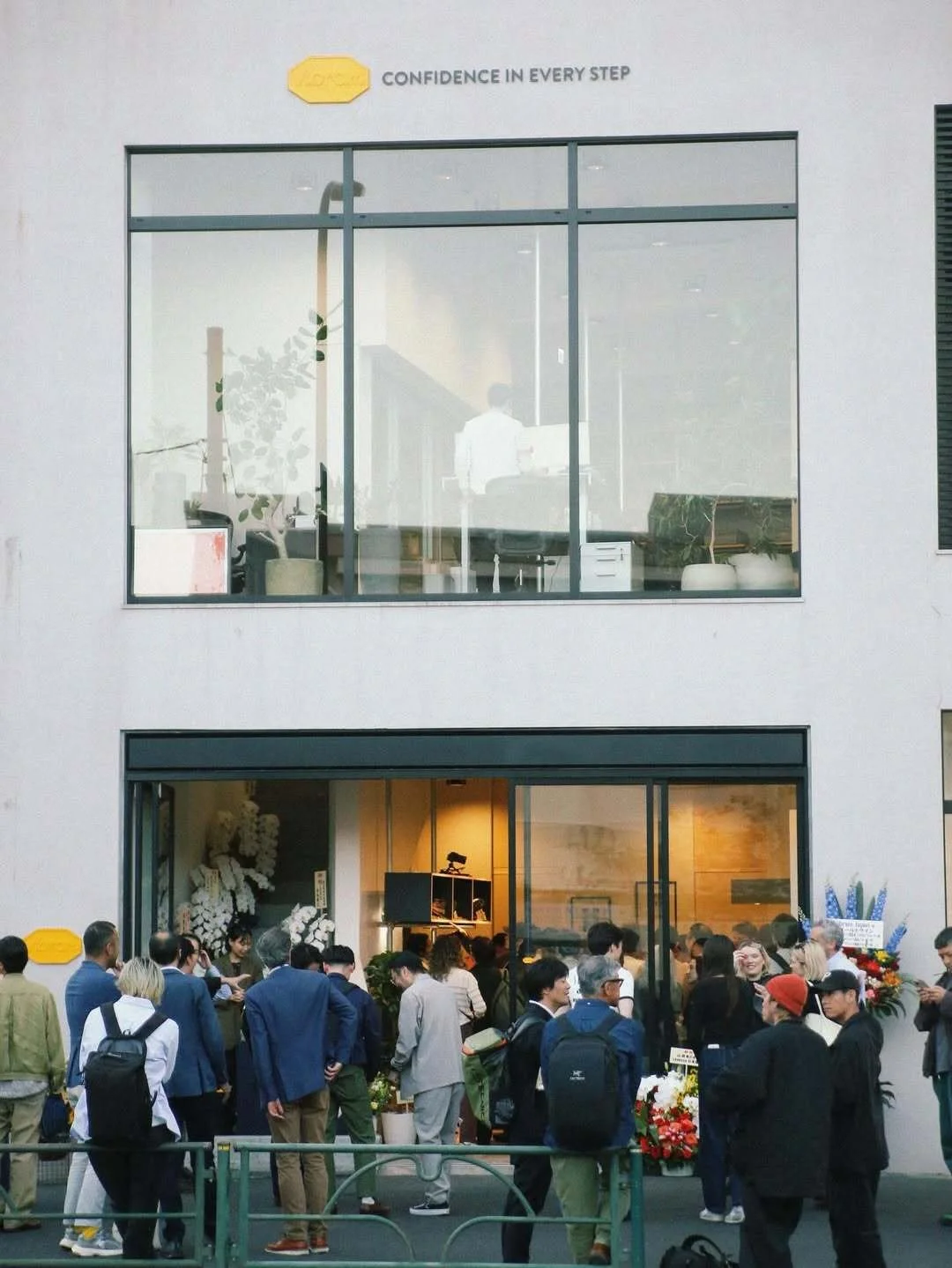 A crowd of people standing outside a storefront with large windows. The store appears to be opening or hosting an event, as many are gathered, some with backpacks. Inside, visible through the glass, is modern decor with plants and awards or plaques. An upper floor with office space and a person working can be seen through the window.
