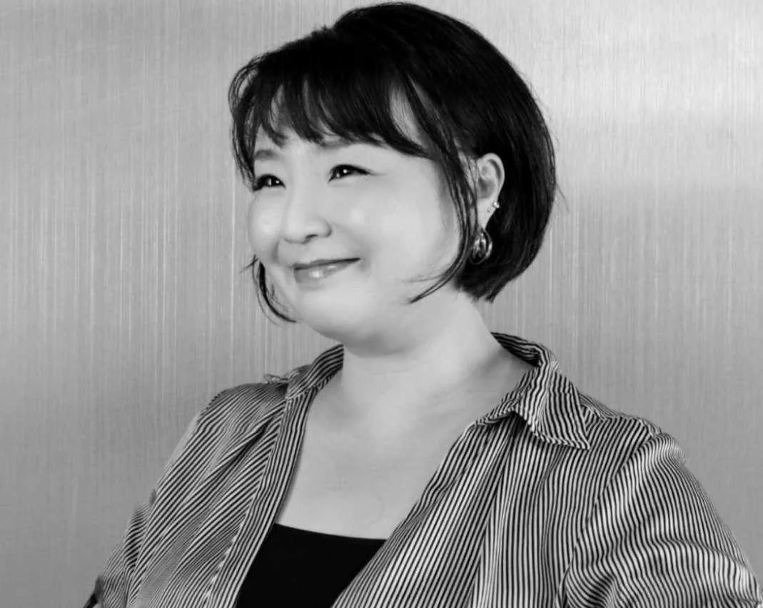 A woman with short dark hair and earrings, smiling, wearing a striped shirt against a plain background.