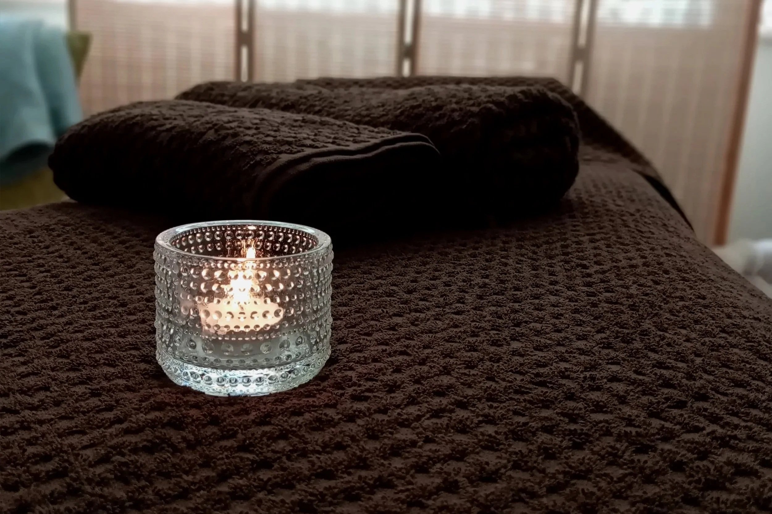 A lit candle in a textured glass holder is placed on a brown, fuzzy surface. In the background, there are three rolled-up brown towels on the same surface and a blurred setting that appears to be a cozy indoor space.