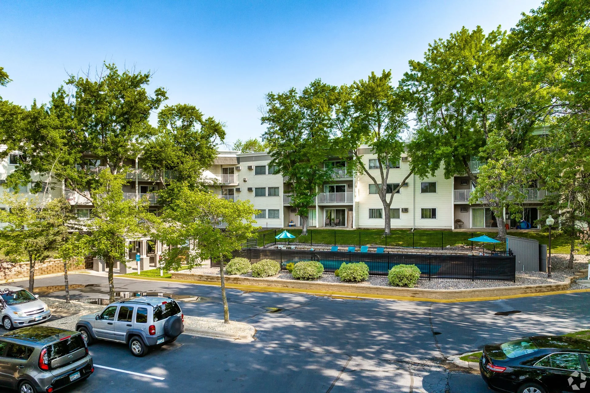 Hillcrest Place apartment complex with trees, a swimming pool, parked cars, and green lawns under a clear blue sky.