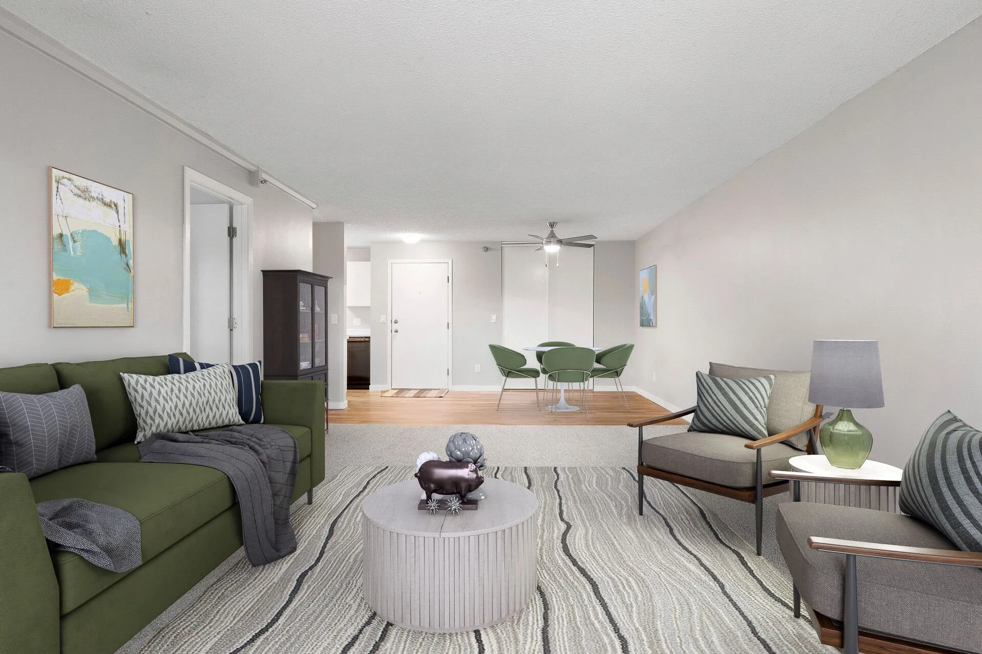 Living room with gray and green furniture, abstract wall art, a white coffee table with decorative sculptures, a dining area with a round white table and green chairs, hardwood and carpeted flooring, and neutral-colored walls.