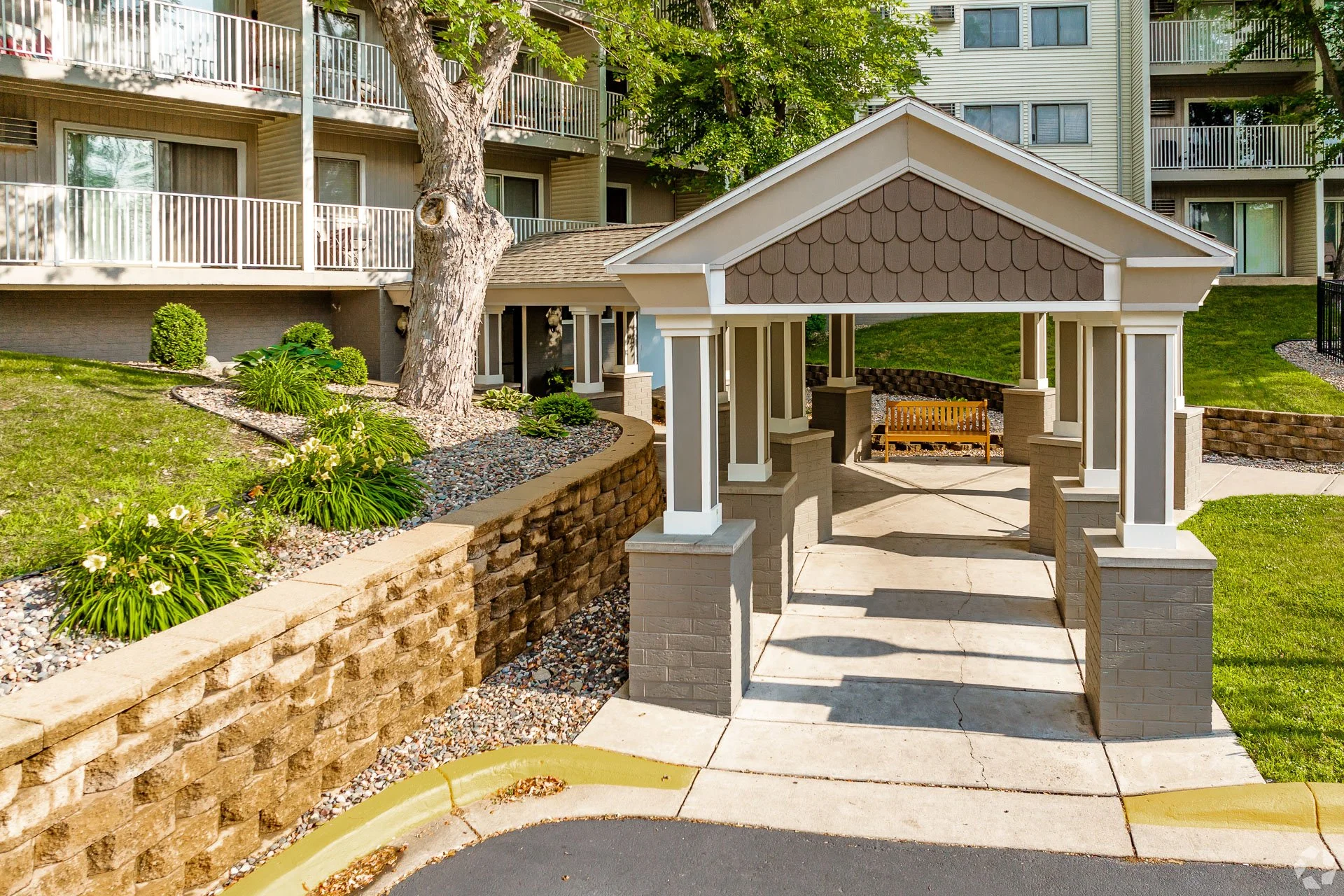 A landscaped apartment complex entrance with a small covered structure, a brick retaining wall, green shrubbery, and a tall tree. Multi-story apartment buildings with balconies are in the background.