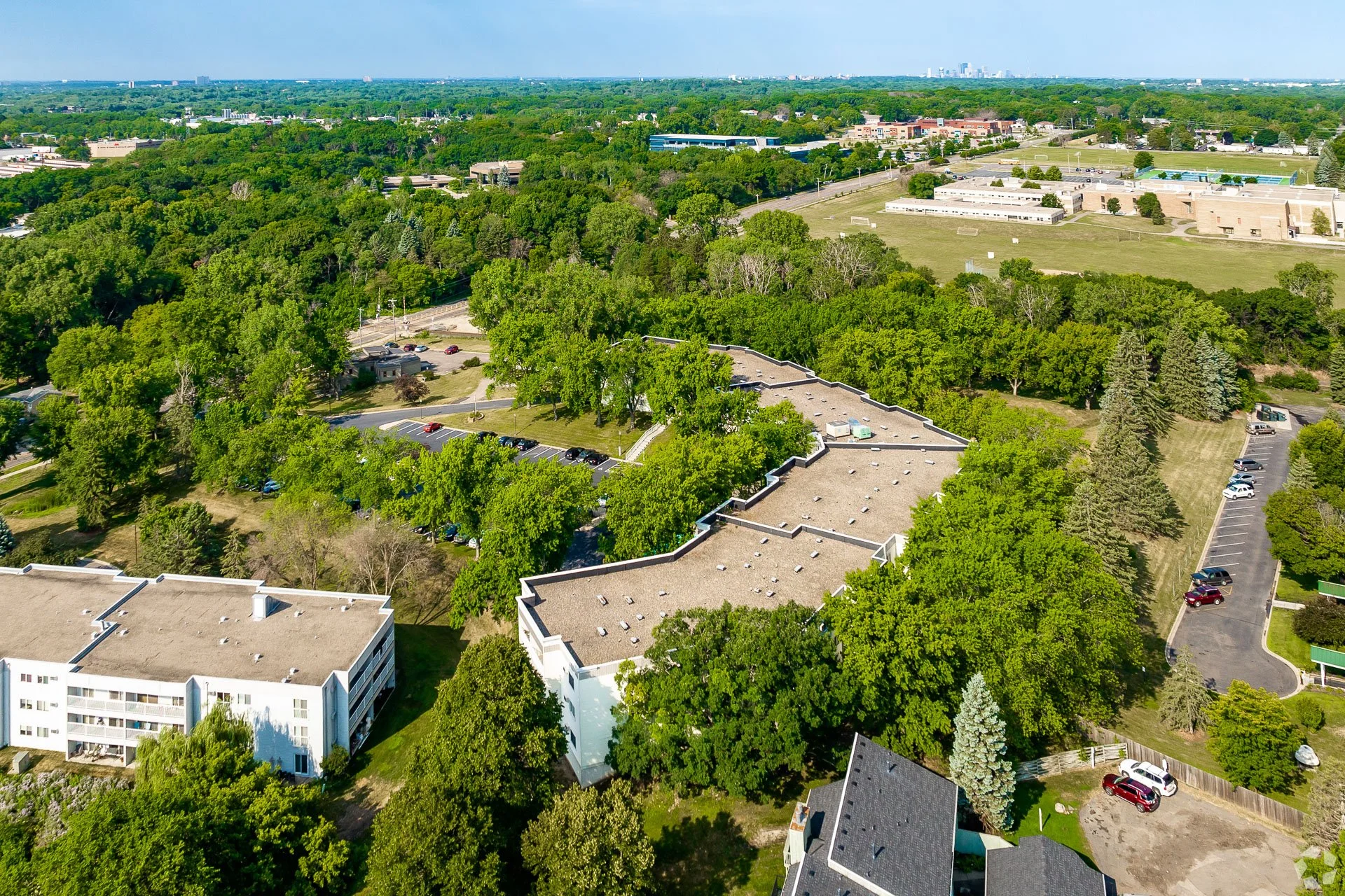 Aerial view of a green suburban area with trees, residential buildings, parking lots, and sports fields with clear blue sky.