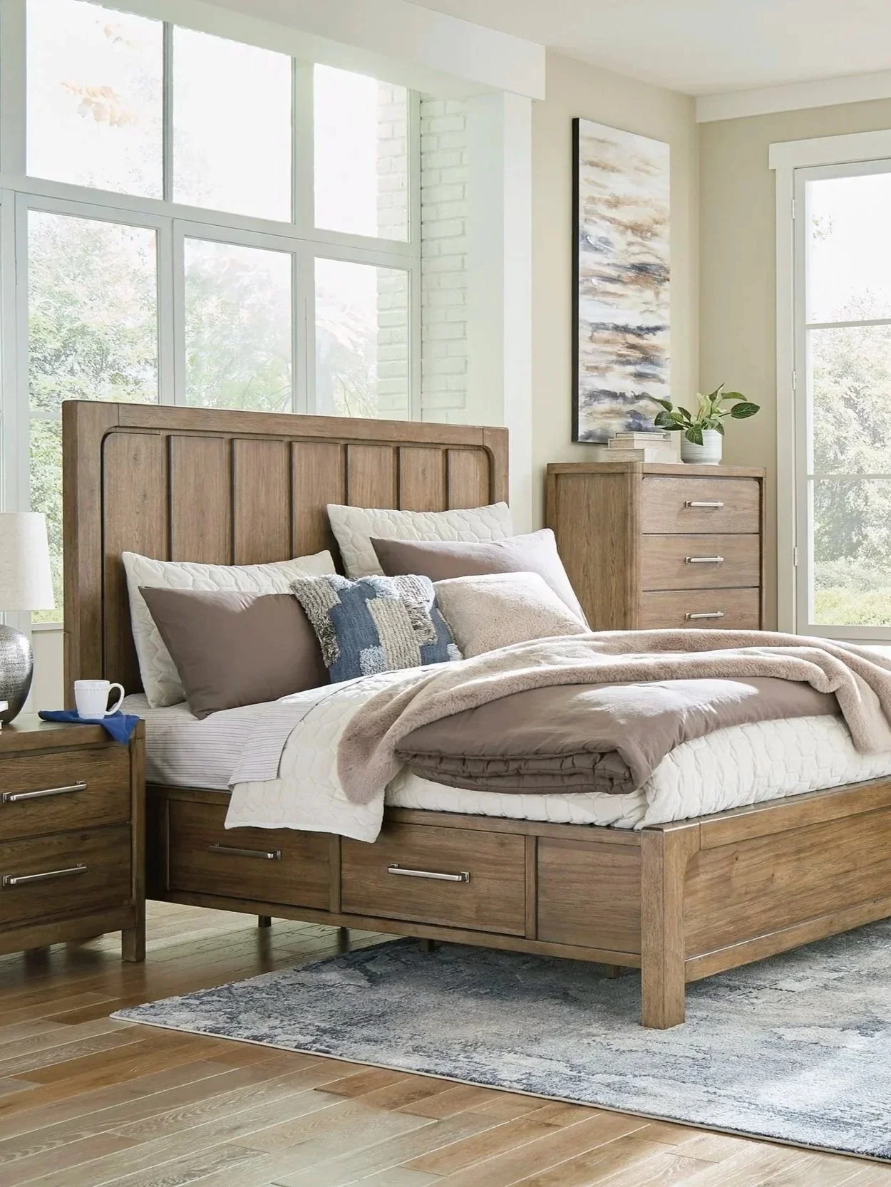 A bedroom with a large wooden bed, neutral-colored bedding, a nightstand with a lamp and mug, and a dresser with books and a potted plant. Large windows let in natural light, and there is a piece of abstract art on the wall.