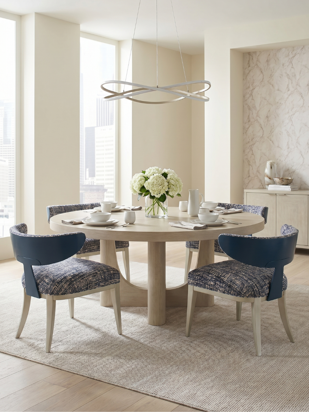 Modern dining room with round wooden table, six chairs with patterned cushions, white vase with white flowers, and minimalist chandelier over the table, large windows in the background.