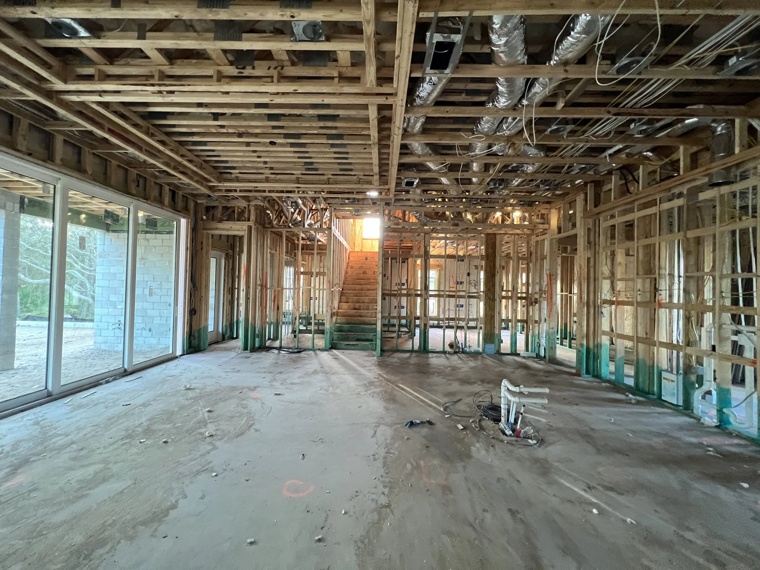 Interior of a house under construction with exposed wooden framing, ductwork, and construction materials on the floor, large windows on the left side, and a staircase towards the back.