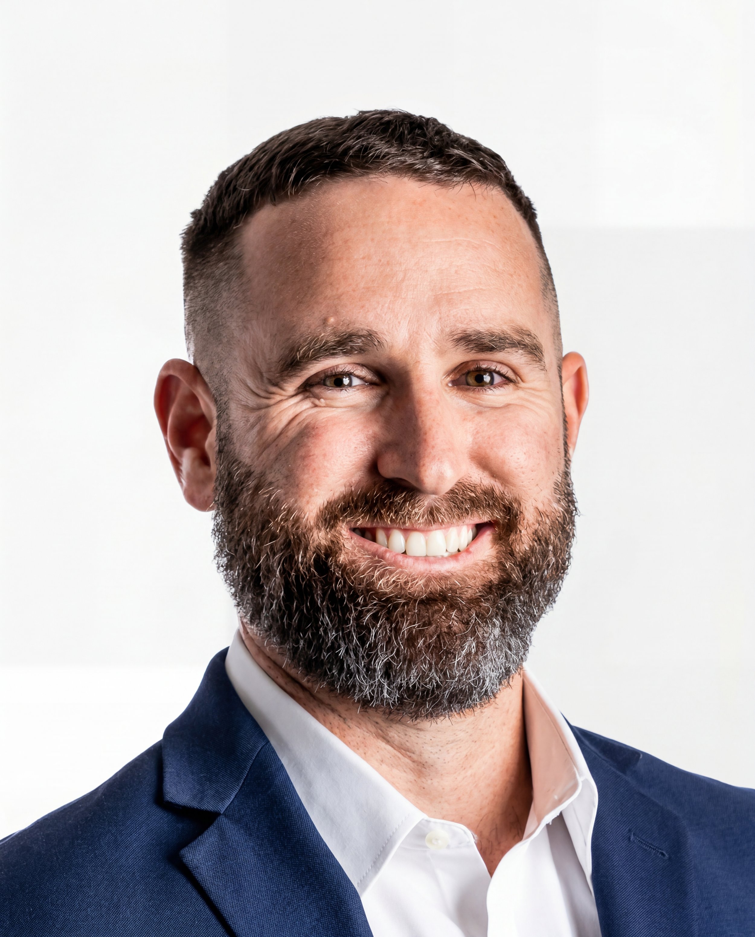 Close-up of a smiling man with a beard dressed in a navy blue suit and white shirt, against a plain light background.