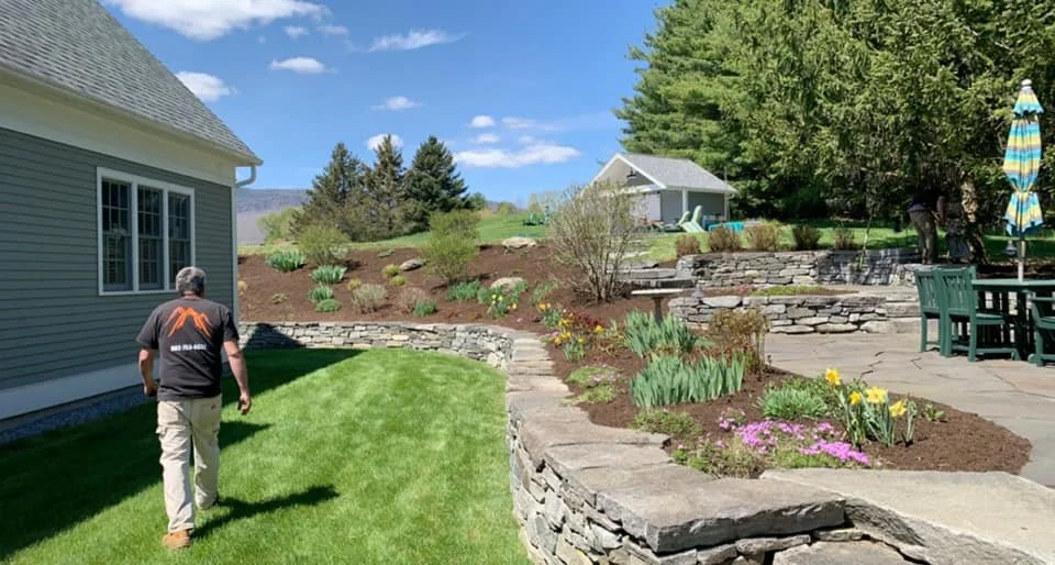 A man in a black shirt and khaki pants walking on a well-maintained lawn next to a house with gray siding. The yard features tiered stone landscaping with blooming flowers and a patio area with outdoor furniture and a striped umbrella. In the background, there are trees, shrubs, and small buildings under a blue sky with clouds.
