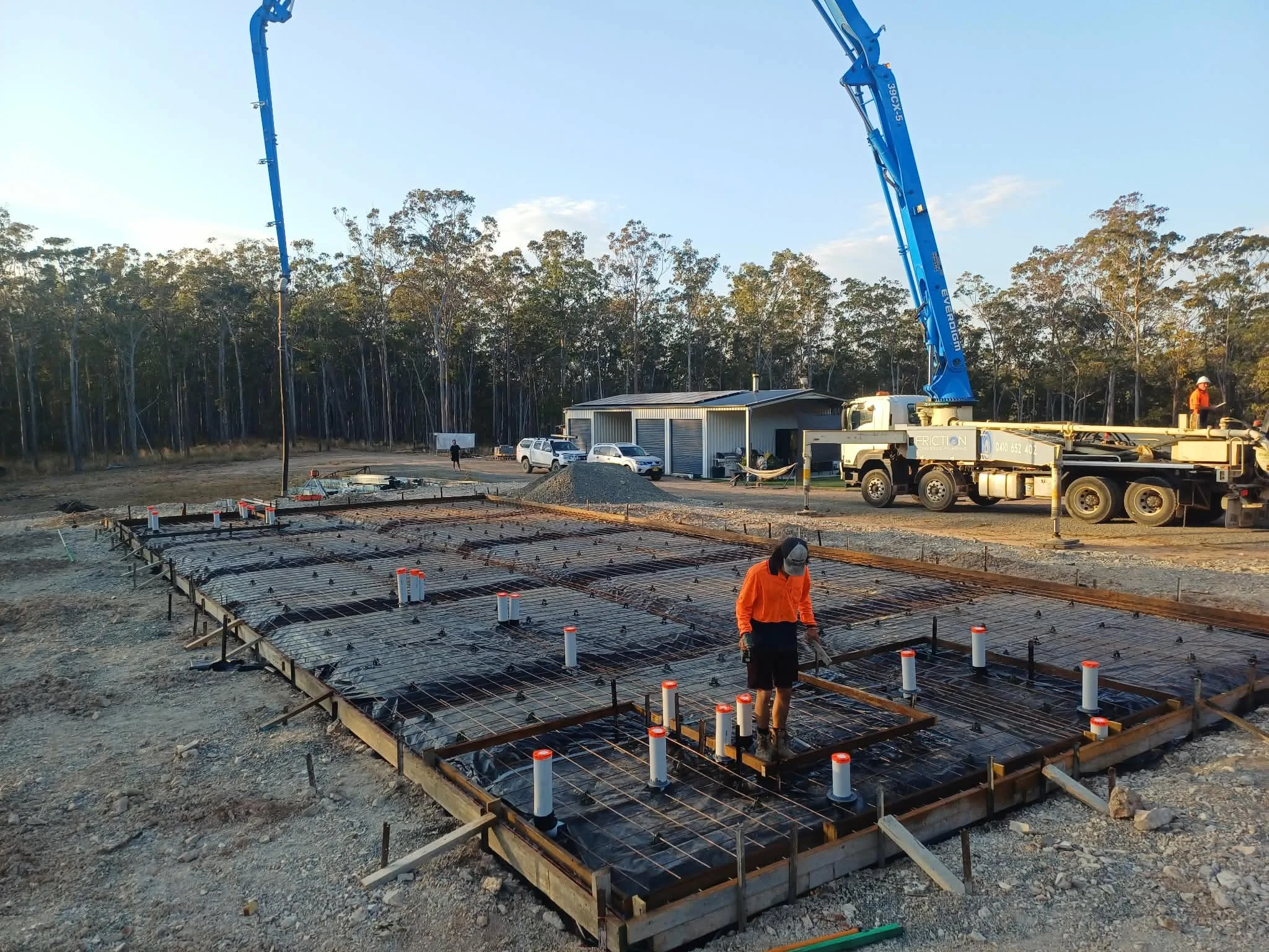 Construction site with metal framework and rebar for a building foundation, a concrete pump truck with a long blue arm, and workers in orange safety vests and hard hats.