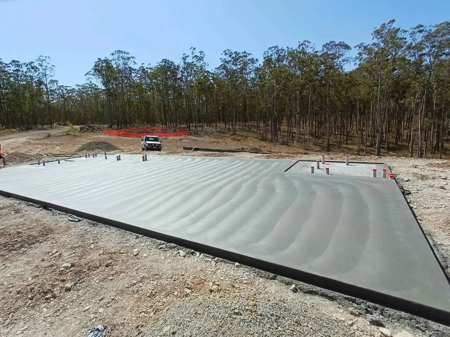 Construction site with a newly laid concrete slab, small pipes protruding, a white truck, and a wooded background.