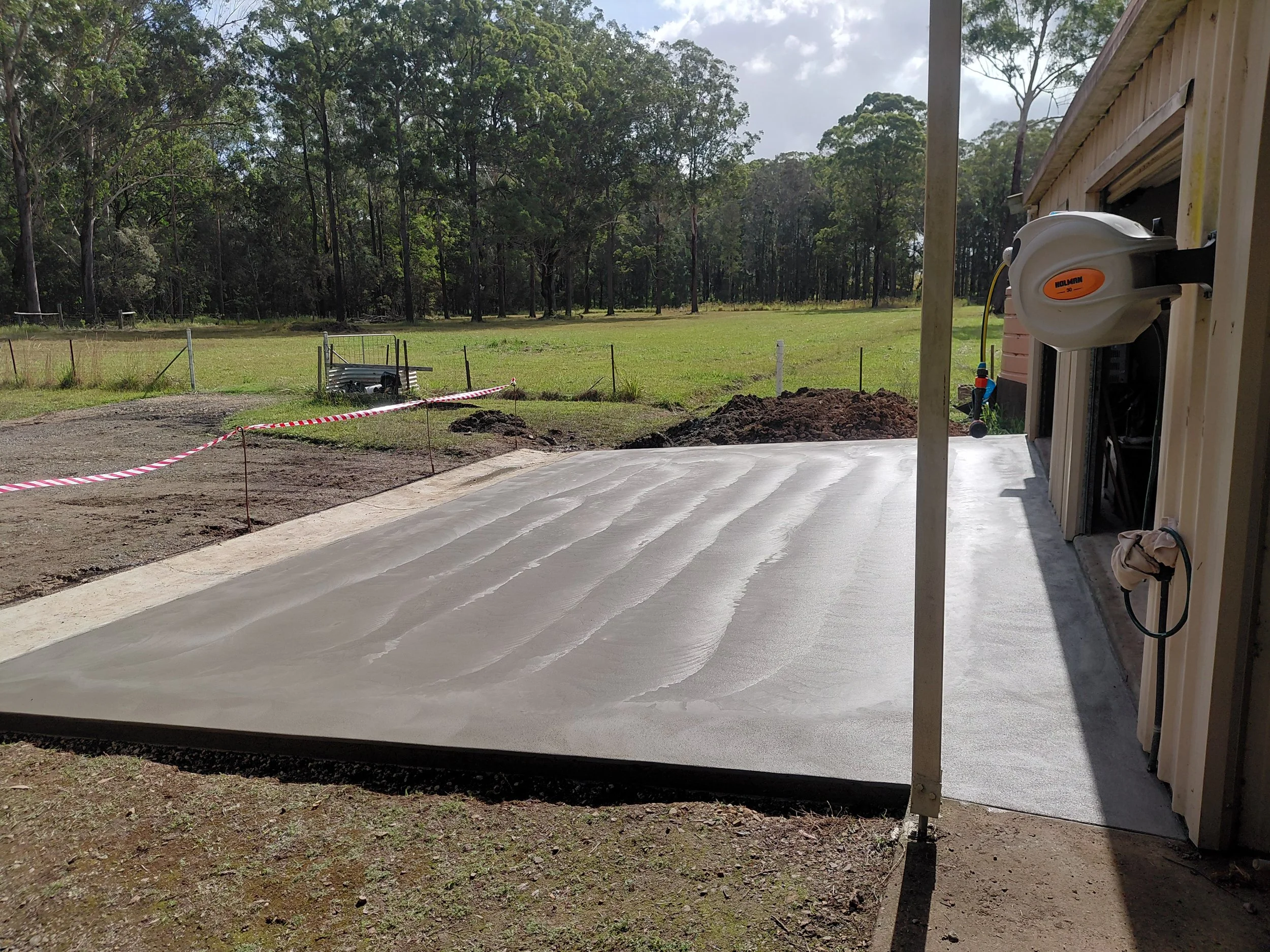 Construction site with freshly poured concrete slab, dirt, and construction materials, with a background of trees and a cloudy sky.