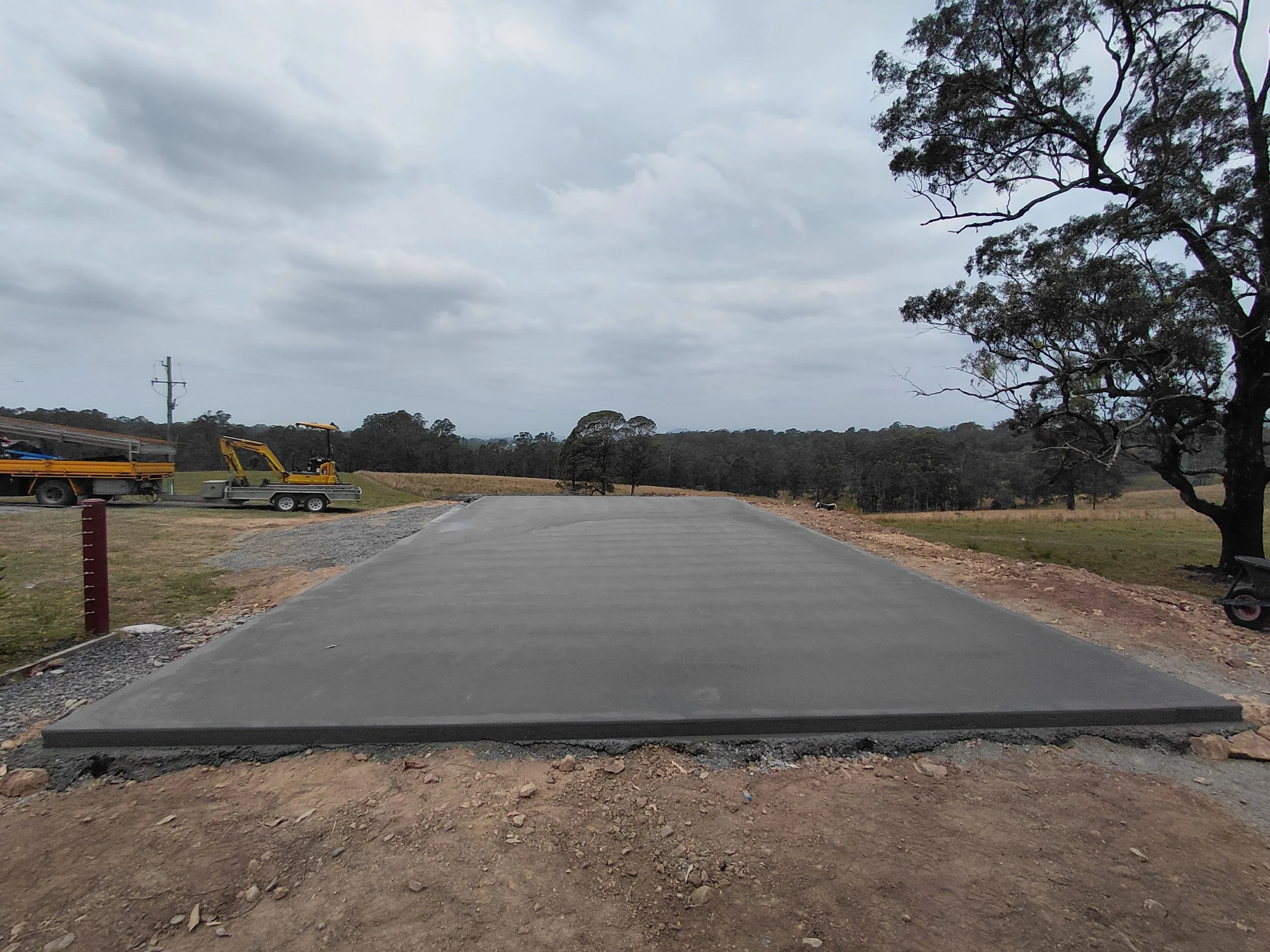Grey overcast sky with some scattered clouds, large trees on the right side of the image, a paved road under construction with a gravel surface ahead, construction equipment on the left, and a utility pole in the distance.