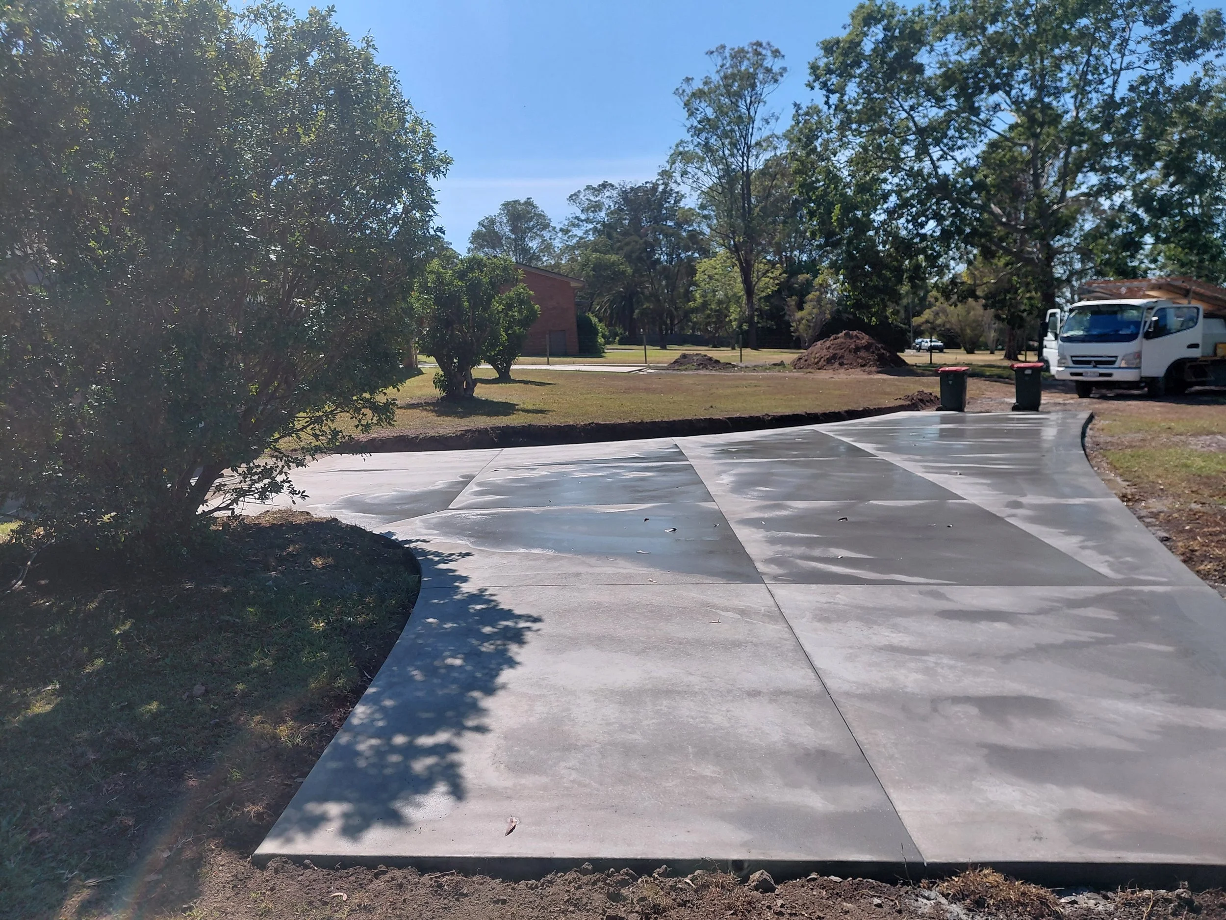 A parking lot with freshly paved concrete, surrounded by green trees and a few small dirt mounds. A white truck is parked on the right side of the image, with two trash bins nearby. A brick building is visible in the background amidst the trees, under a clear blue sky.