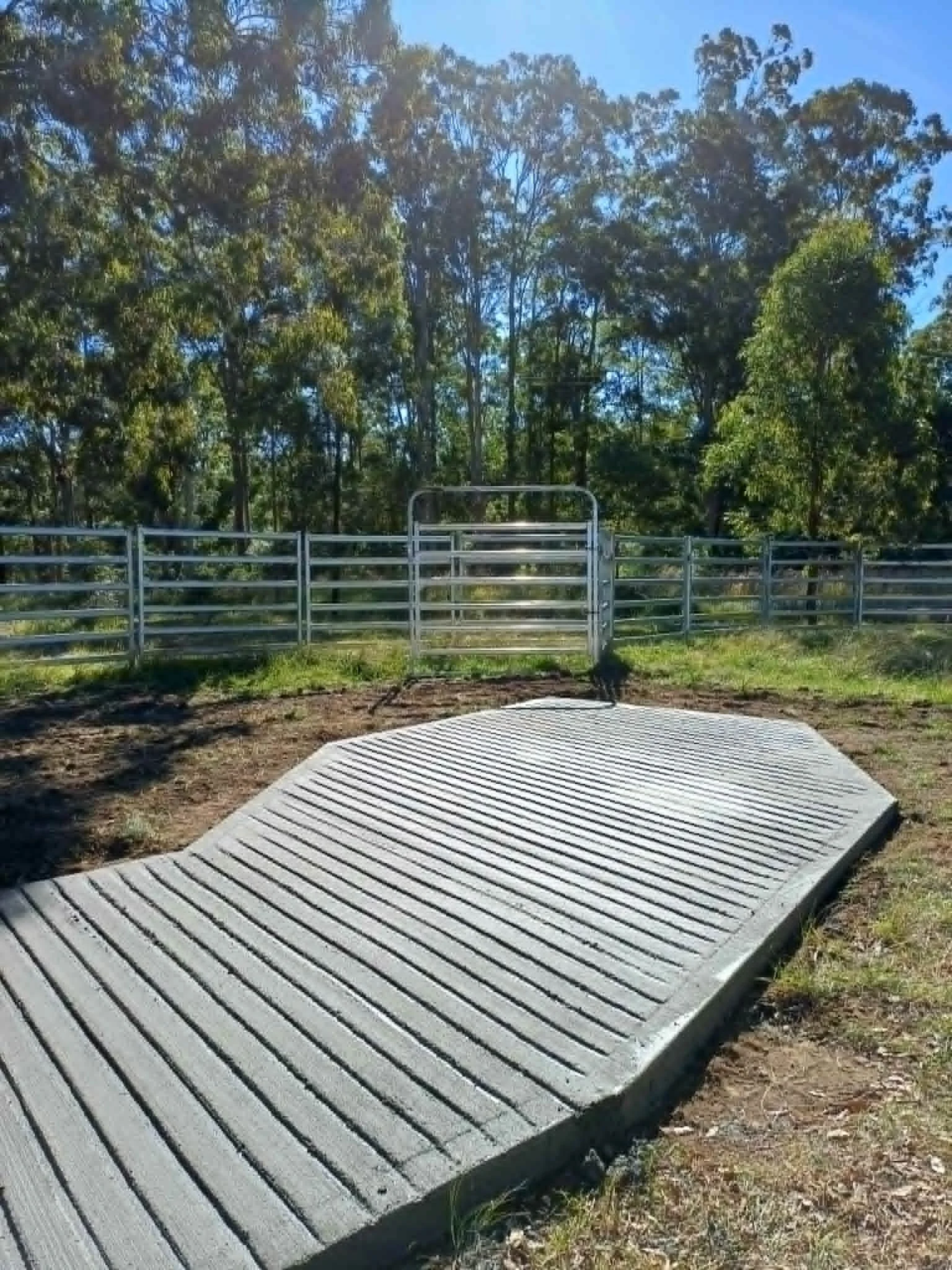 A metal cowling mat placed on grassy ground with a metal gate and fence behind it, and a backdrop of tall trees and blue sky.