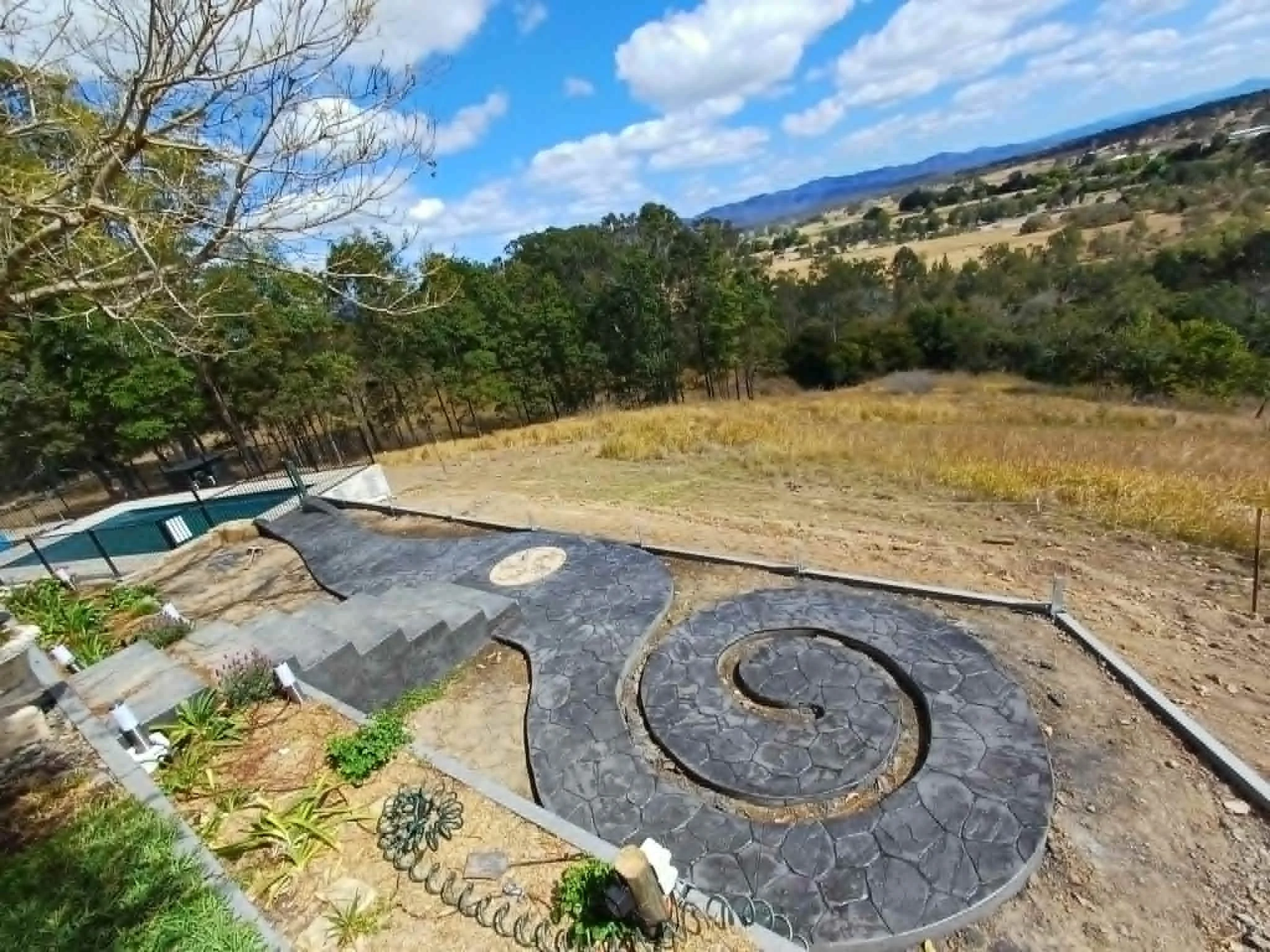 A scenic landscape with trees, mountains in the distance, and a partly cloudy sky. Includes a garden area with plants and a small trampoline, with a gravel or dirt path.