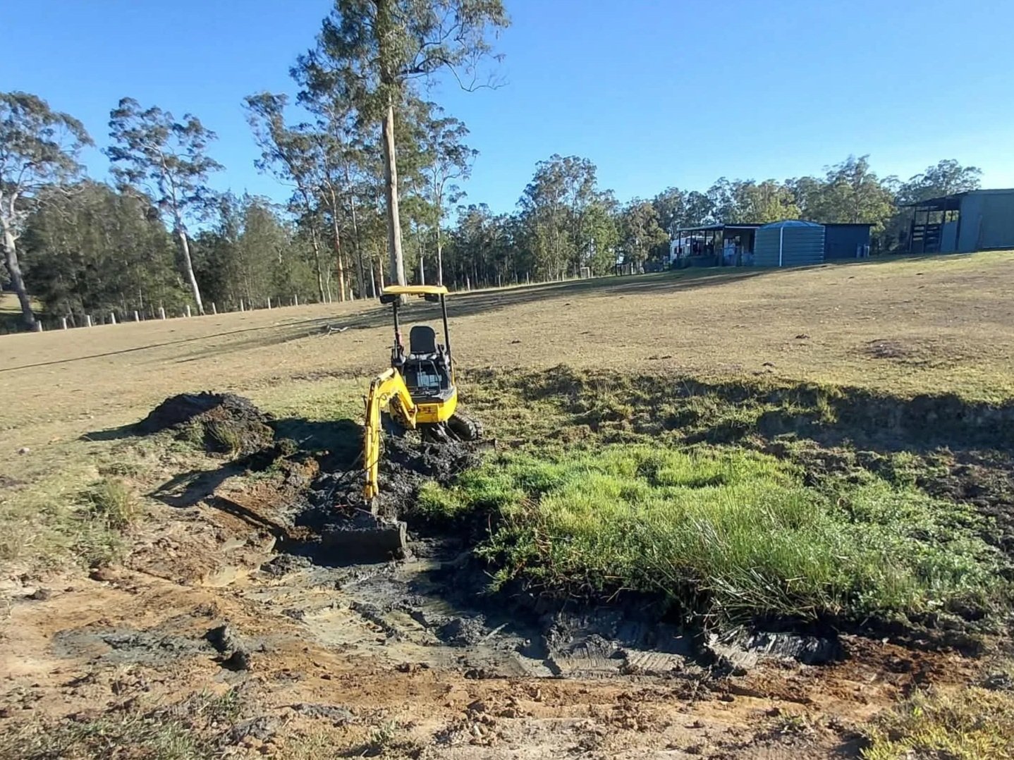 A small yellow excavator working on a construction site with grass and dirt in a rural area, with trees and a few buildings in the background.