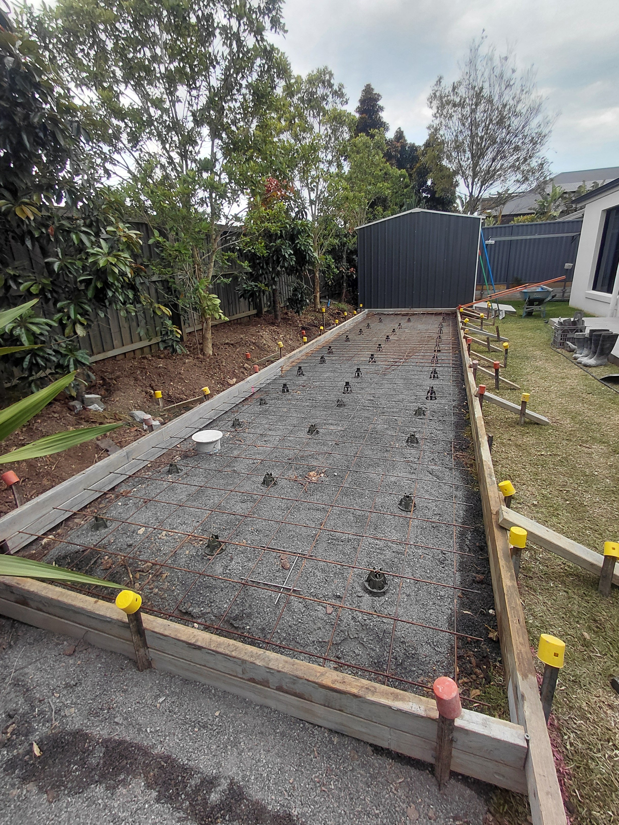A construction site showing a wooden frame with rebar and orange string, likely for a concrete slab, next to a garden with trees and shrubs, and a blue shed in the background.