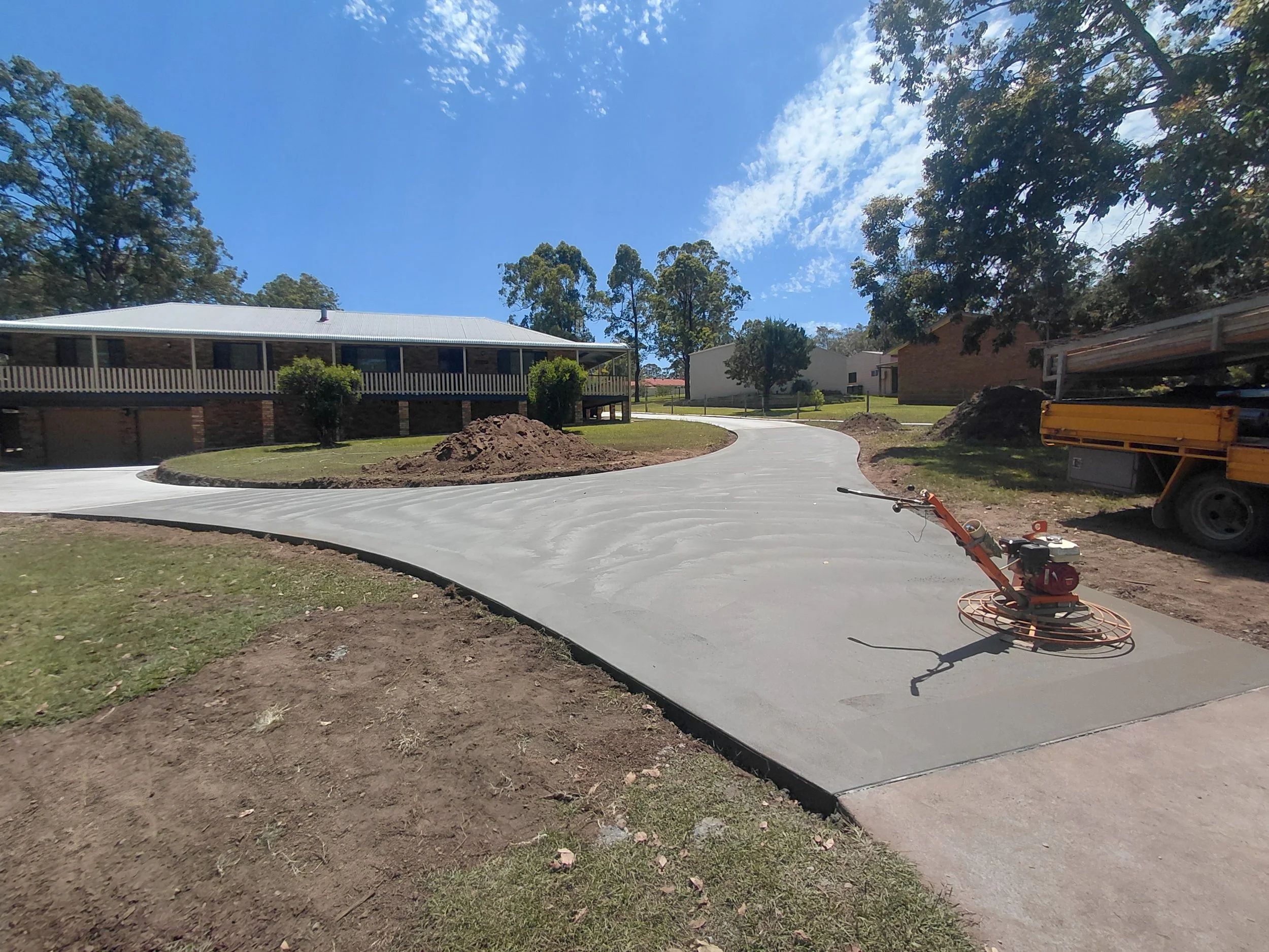 Residential construction site with newly poured concrete driveway, construction equipment, and houses in the background under a partly cloudy sky.