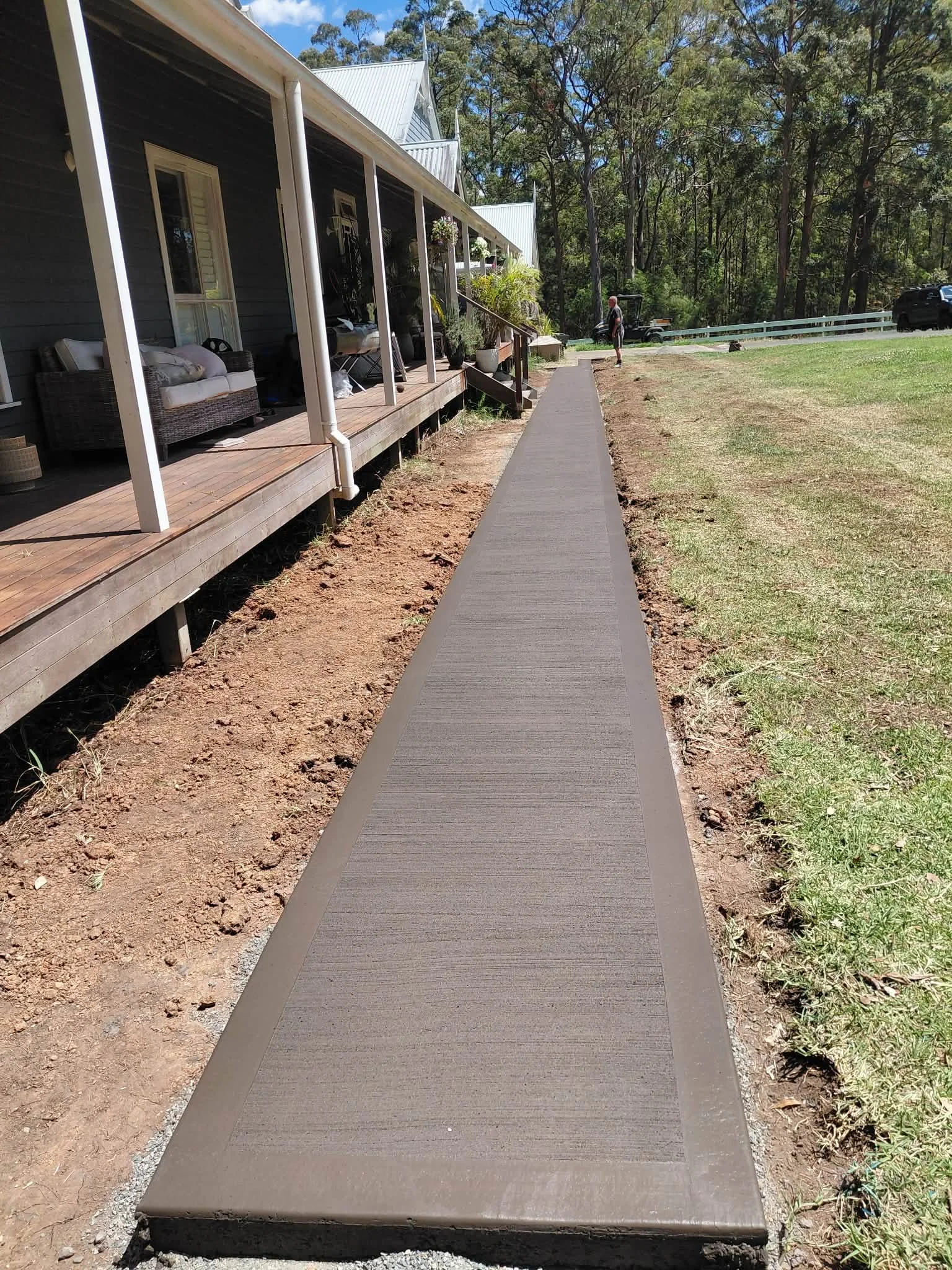 A new concrete sidewalk next to a house with a porch, with a person walking in the background in a grassy area surrounded by trees.