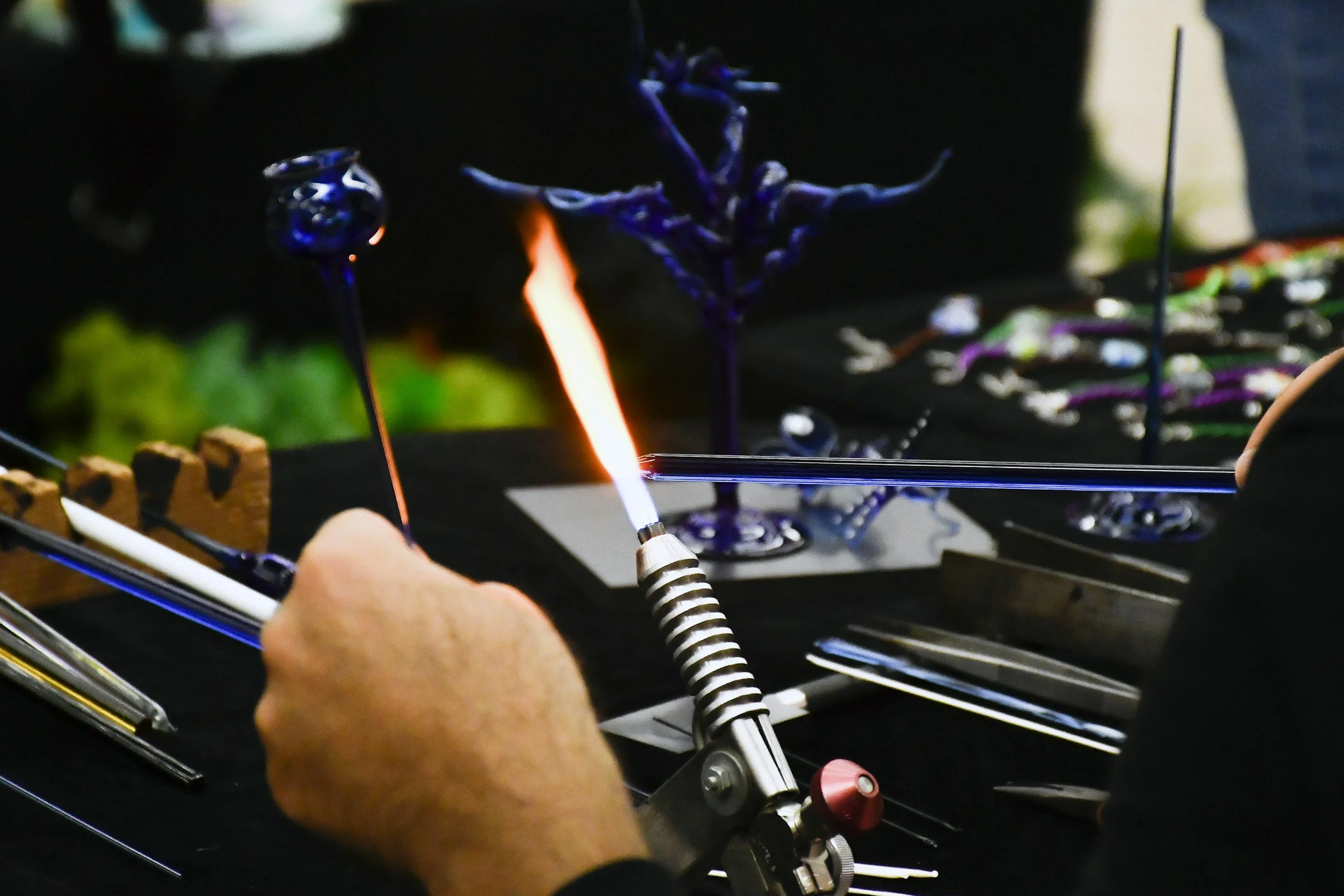 Manos manipulando vidrio fundido con un sople de gas, en un taller de vidrio artesanal, con figuras de vidrio en el fondo.