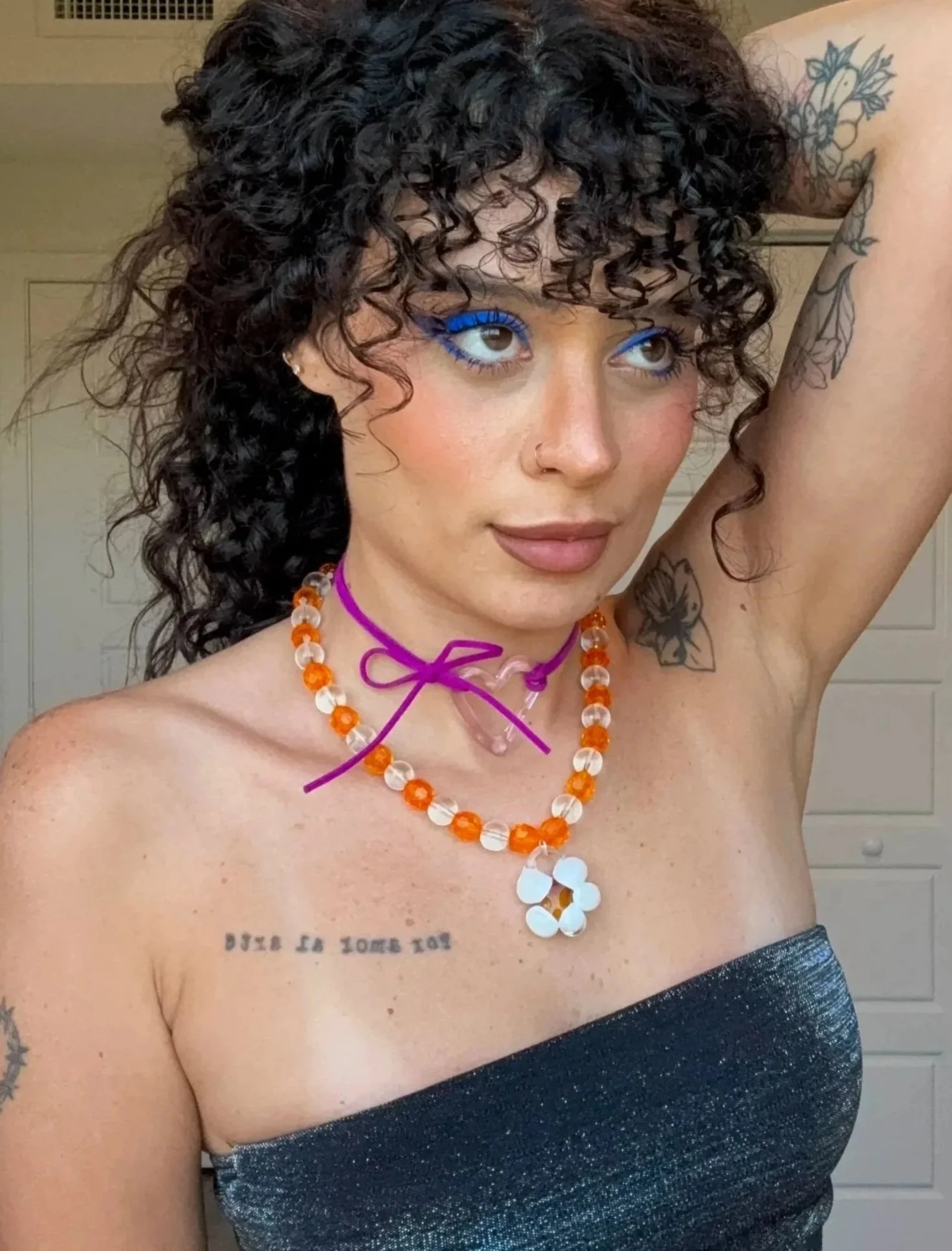 Mujer con cabello rizado y oscuro, que lleva joyas en el cuello, maquillaje llamativo en los ojos, y tatuajes en los brazos, con un vestido de tirantes oscuro y brillo.
