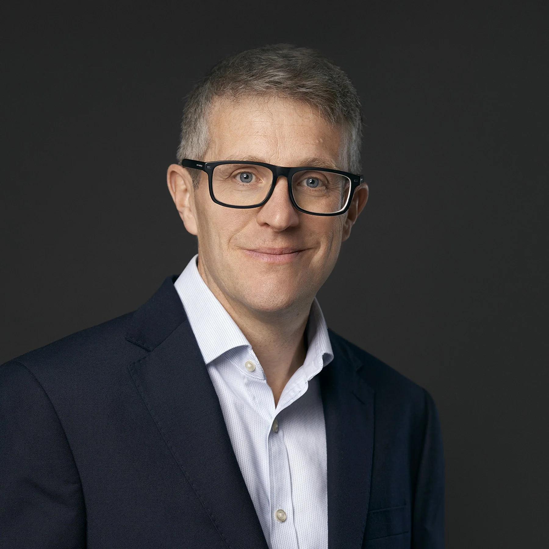 Close-up of a middle-aged man with short gray hair, wearing black rectangular glasses, a white dress shirt, and a dark blazer against a black background.