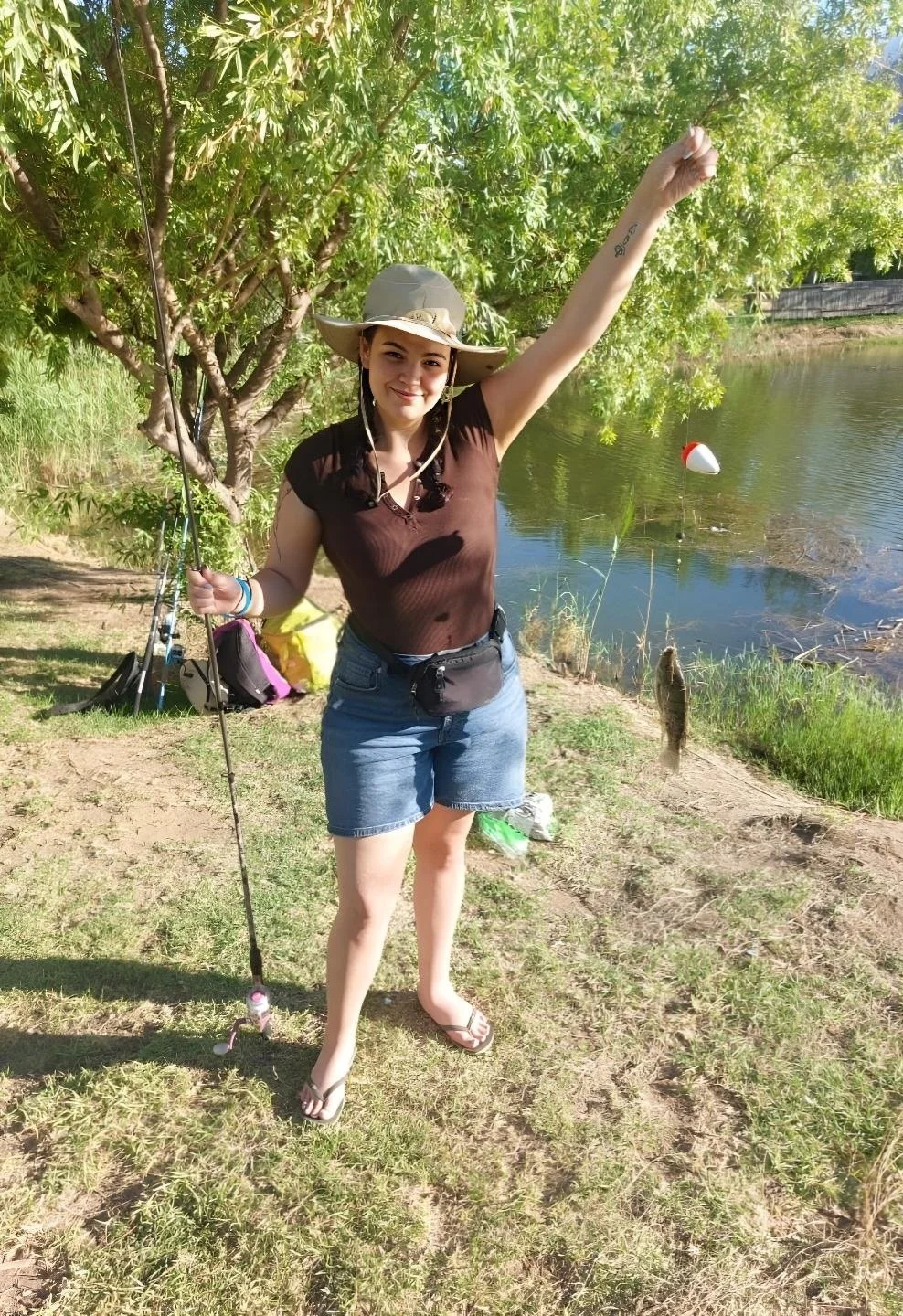 A young woman stands by a lake holding a fishing rod with a fish caught on the line, smiling and raising her arm in triumph, wearing a wide-brimmed hat, a brown shirt, and denim shorts.