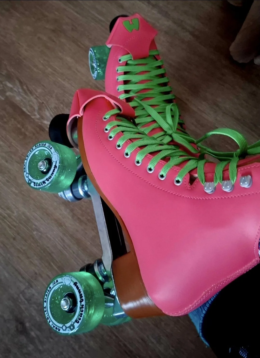 Pink roller skates with green laces and green wheels on a wooden floor.