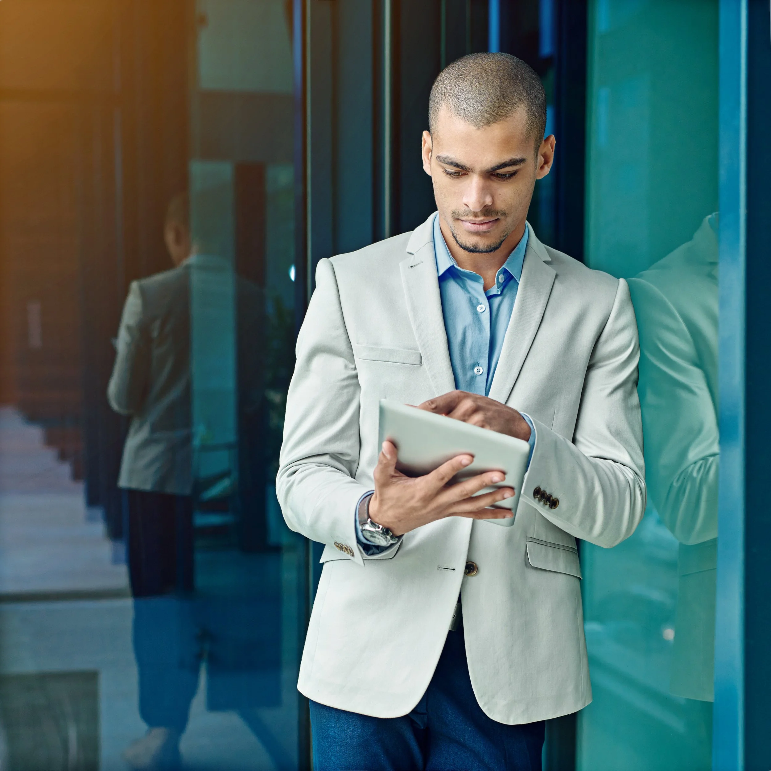 Young man with short hair, wearing a light grey blazer and blue shirt, standing in front of a reflective glass wall, looking at his phone.