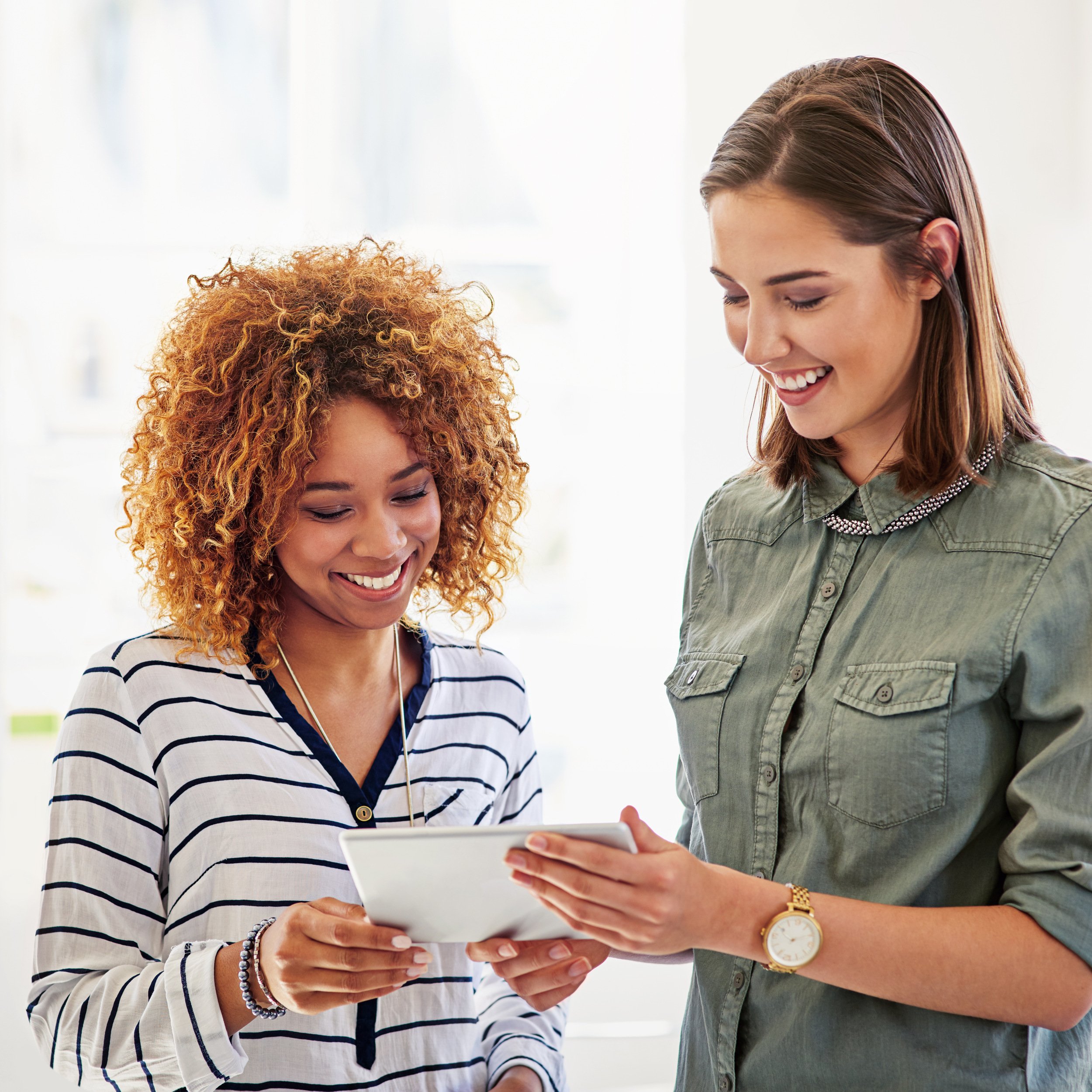 Two women standing together looking at a tablet, smiling, in a bright room with white background.