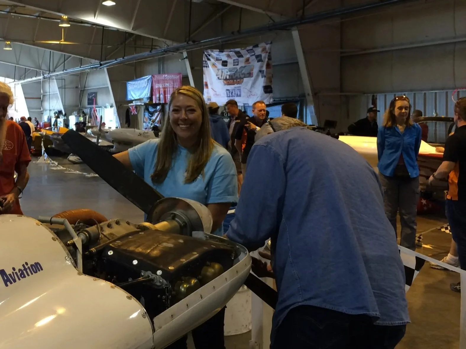 Smiling woman working on a small aircraft inside an airplane hangar, with other people and aircraft in the background.