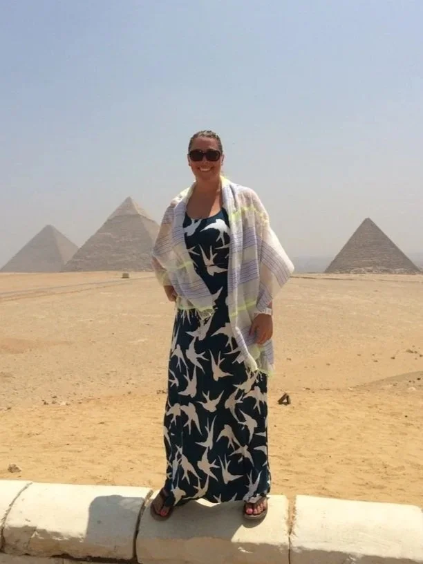 A woman in sunglasses and a flowy dress standing on a sidewalk in front of the pyramids of Giza in Egypt.