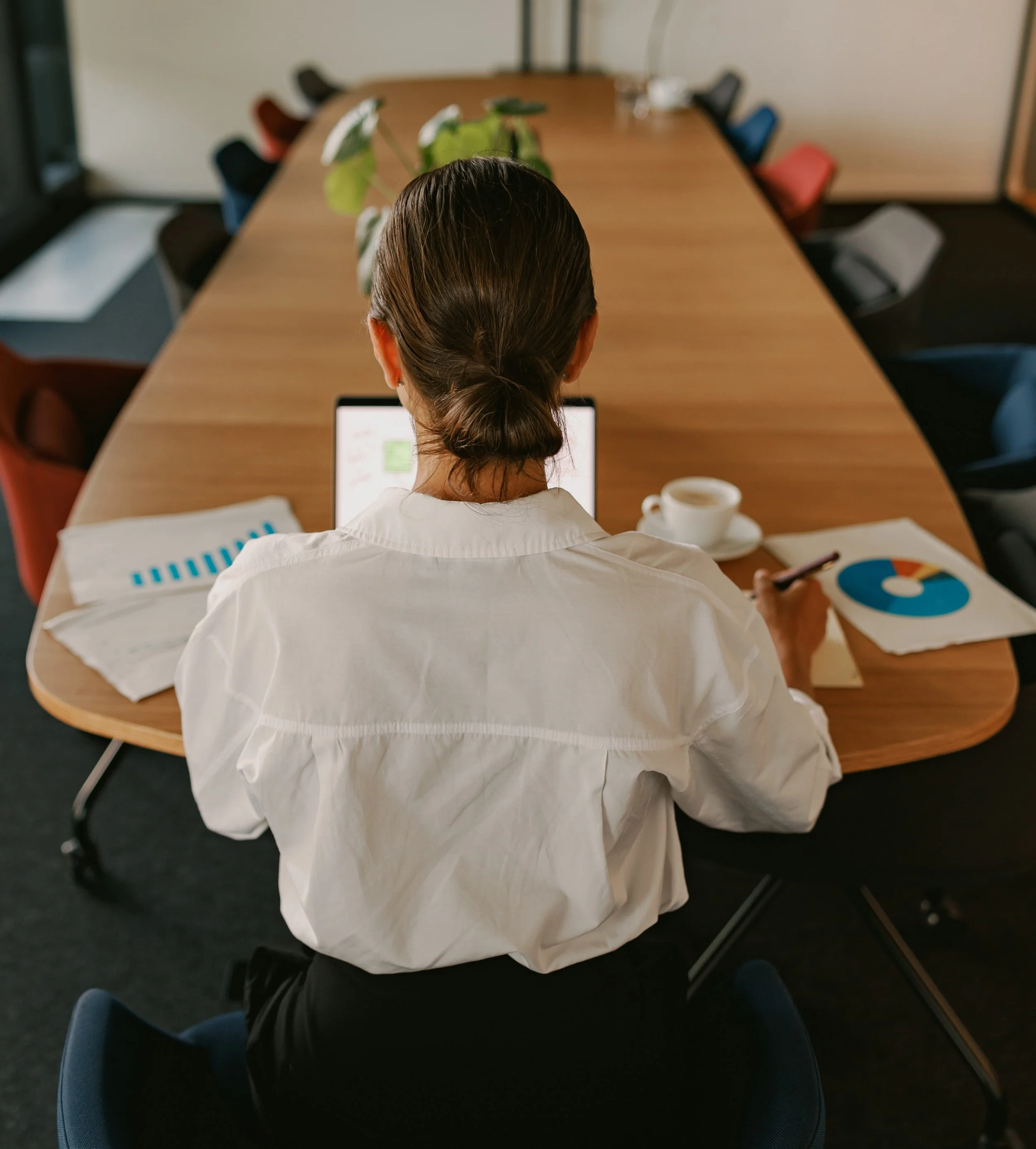 Top view of a woman with brown hair in a ponytail working on a laptop at a conference table with documents, a cup of coffee, and a smartphone nearby.
