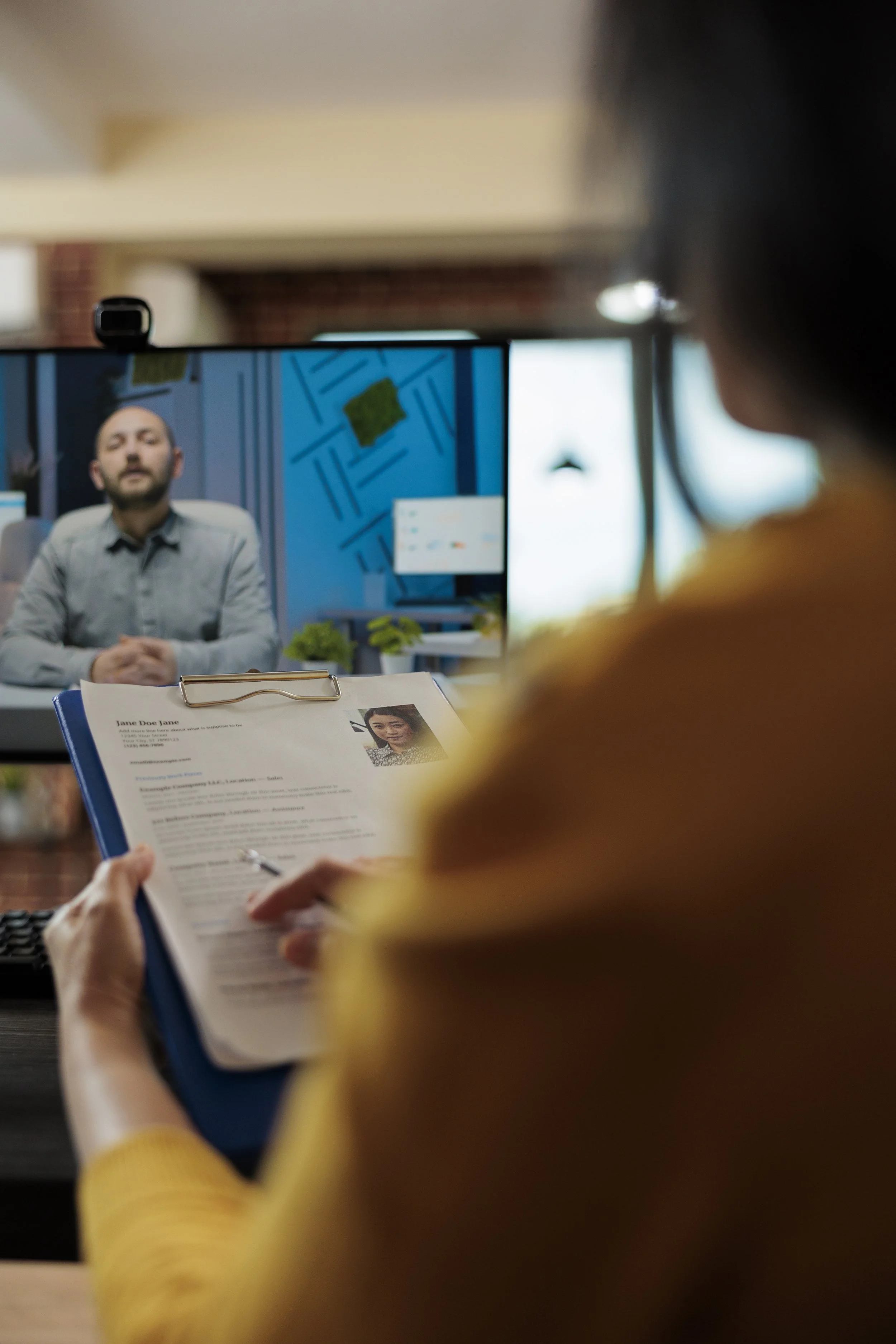 A person in a yellow sweater reviewing a resume with a photo and name, seated at a desk, with a computer monitor displaying a man in an office background in front of him.