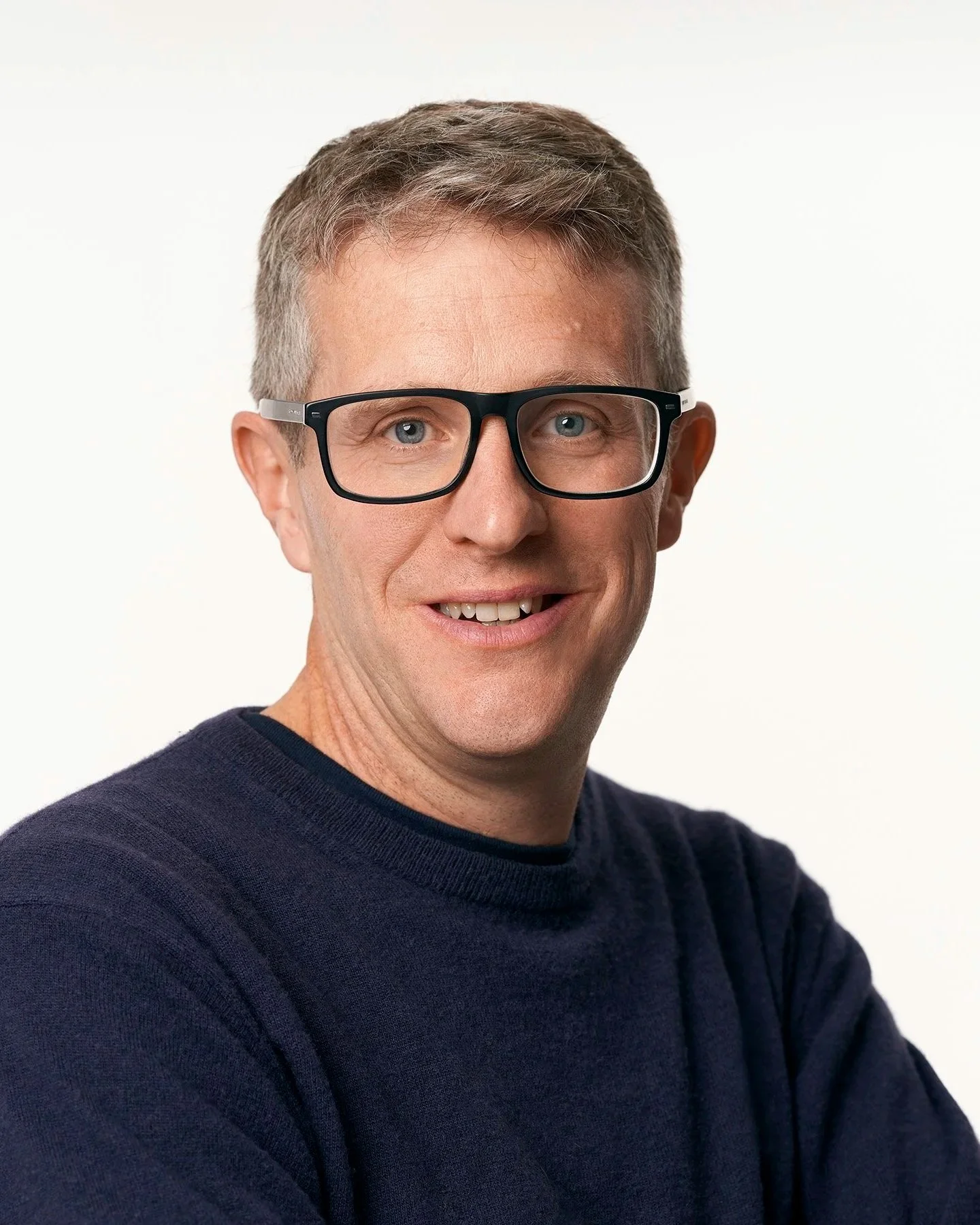 Headshot of a middle-aged man with light skin, short gray hair, blue eyes, wearing black glasses and a navy blue sweater, smiling against a plain white background.