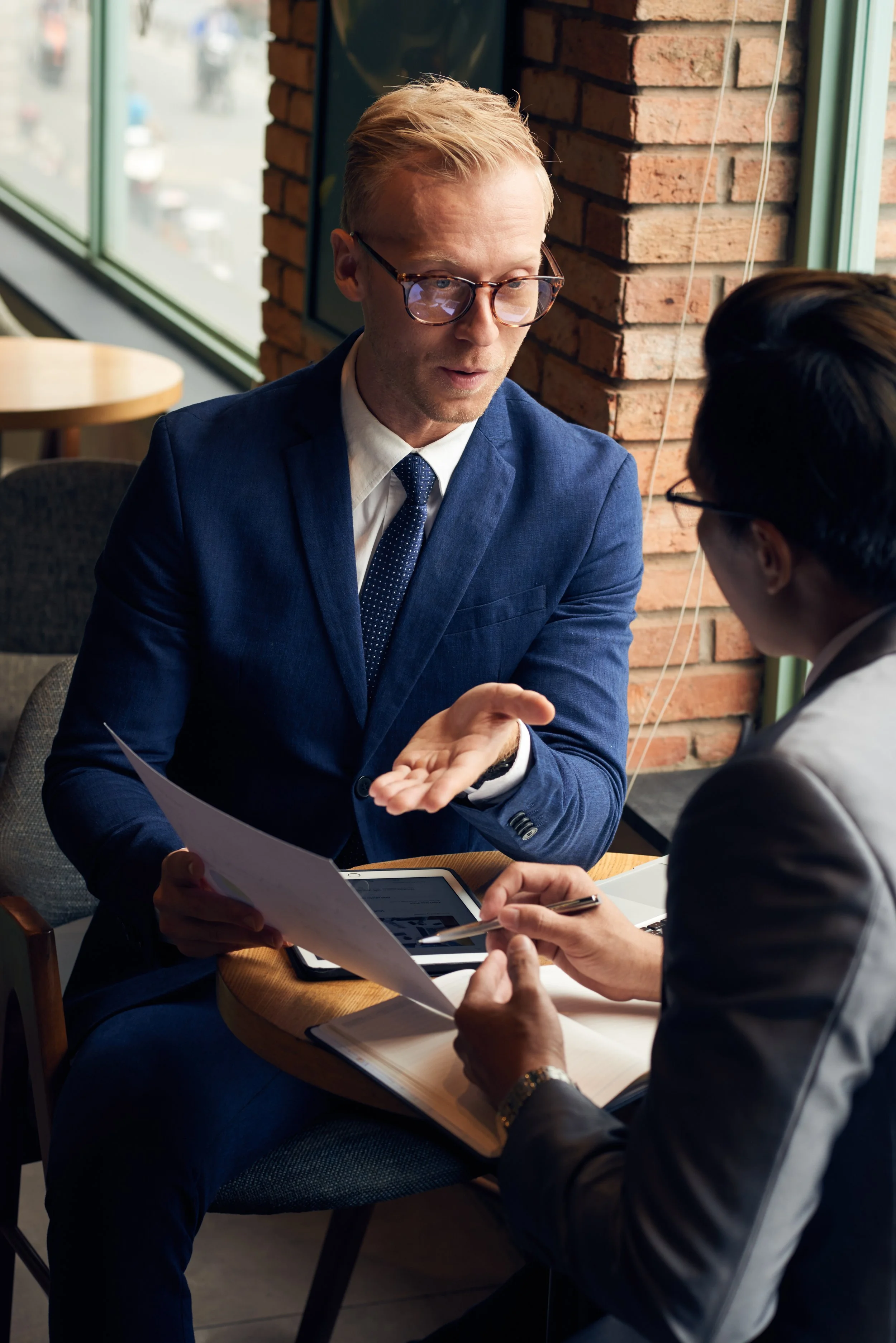 Two men in business suits having a discussion at a table in a cafe, with documents and a tablet in front of them, near a large window and brick wall.