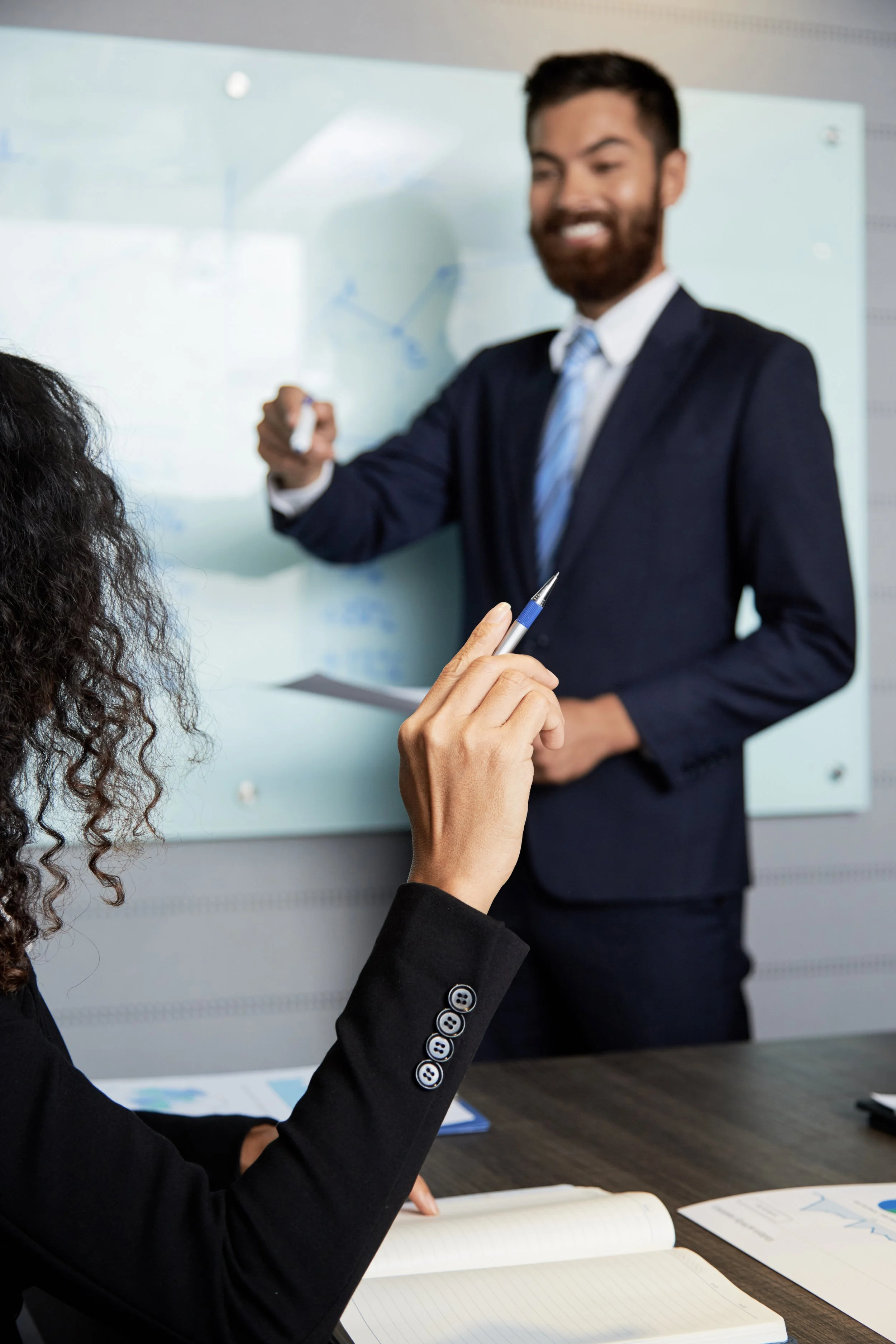 A professional man in a suit is standing in front of a whiteboard, smiling while giving a presentation. A woman seated at the table is taking notes with a pen.