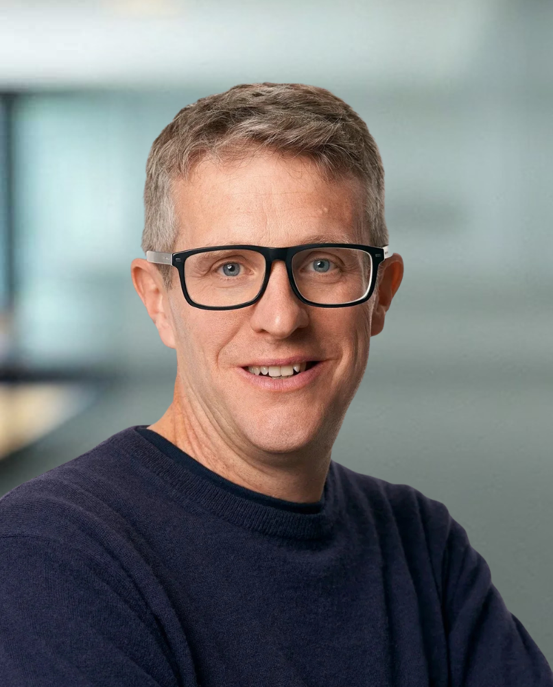 A middle-aged man with short, light brown hair, wearing black-rimmed glasses, smiling, dressed in a dark blue sweater, against a plain white background.
