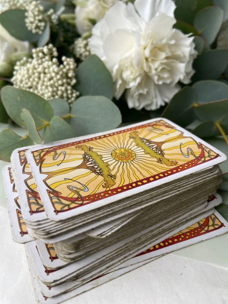 Un tas de cartes de tarot empilées sur une surface blanche, avec des fleurs blanches et des feuilles vertes en arrière-plan.