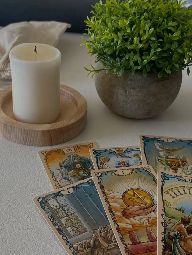 Des cartes de tarot sont étalées sur une table, accompagnées d'une plante verte en pot, d'une bougie blanche dans un porte-b quelle en bois, et d'un coussin blanc en arrière-plan.