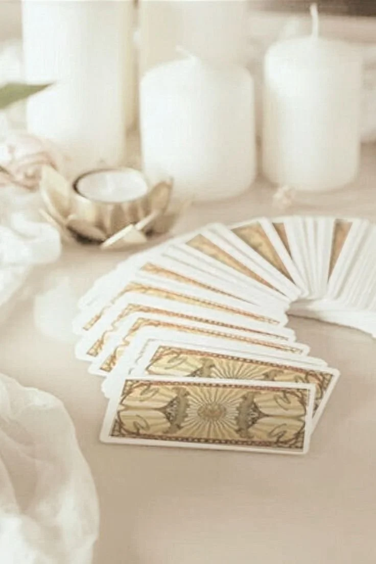 Des cartes de tarot ou de jeu de cartes, disposées en éventail sur une surface claire, avec des bouteilles et des objets blancs en arrière-plan.
