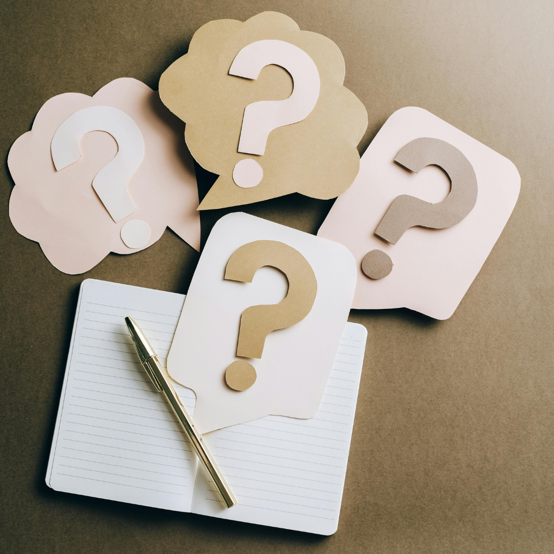 Multiple paper clouds and note cards with question marks, a notebook, and a pen on a brown surface.