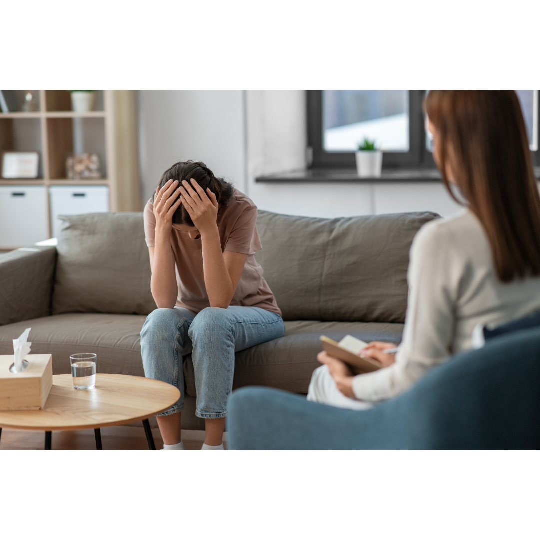 A woman sitting on a couch with her head in her hands, appearing distressed, in a therapy session with a therapist taking notes, in a modern living room.