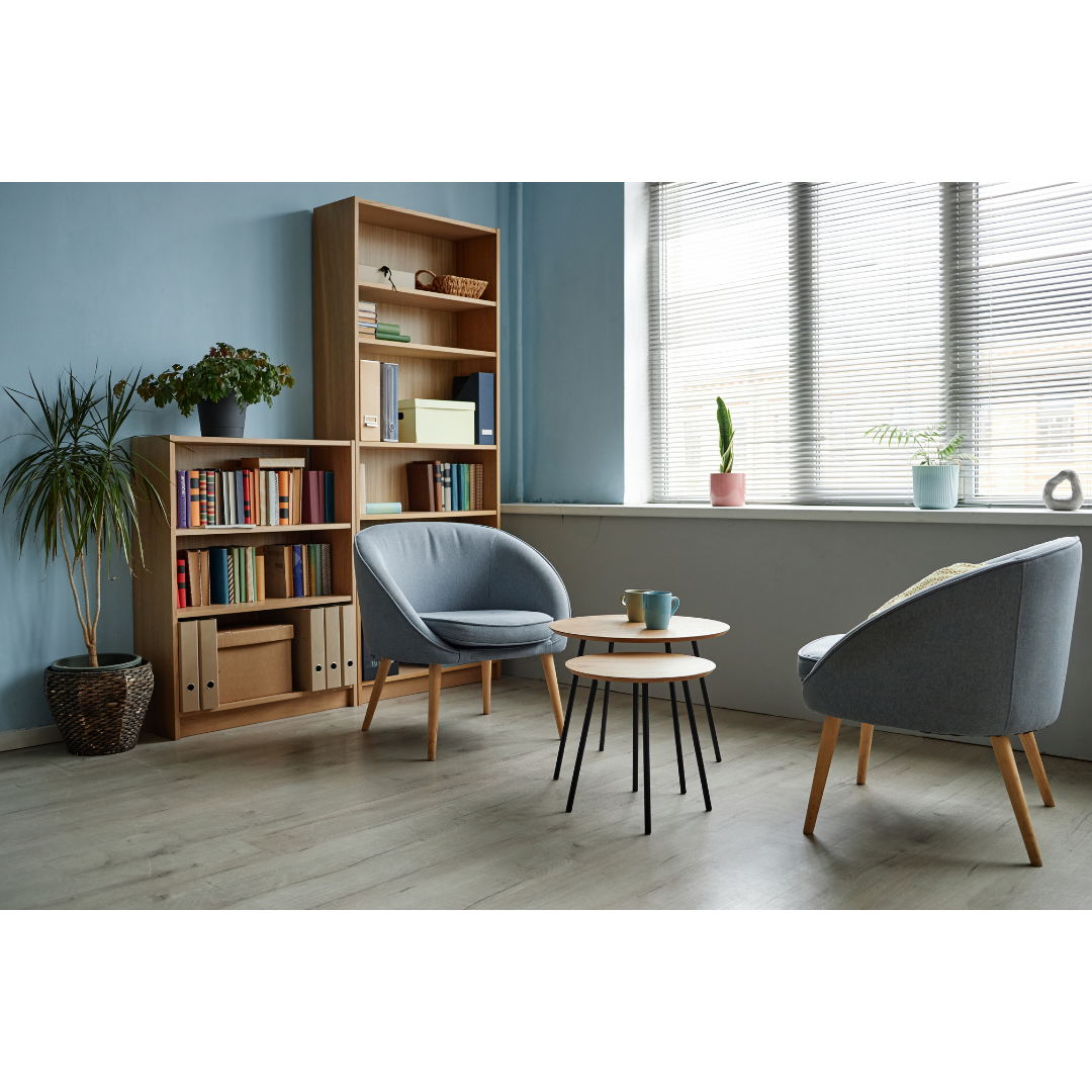 A cozy reading nook with two gray armchairs and a set of small round wooden tables with cups. Behind, a large window with blinds, decorated with potted plants. To the left, a wooden bookshelf and a tall potted plant.
