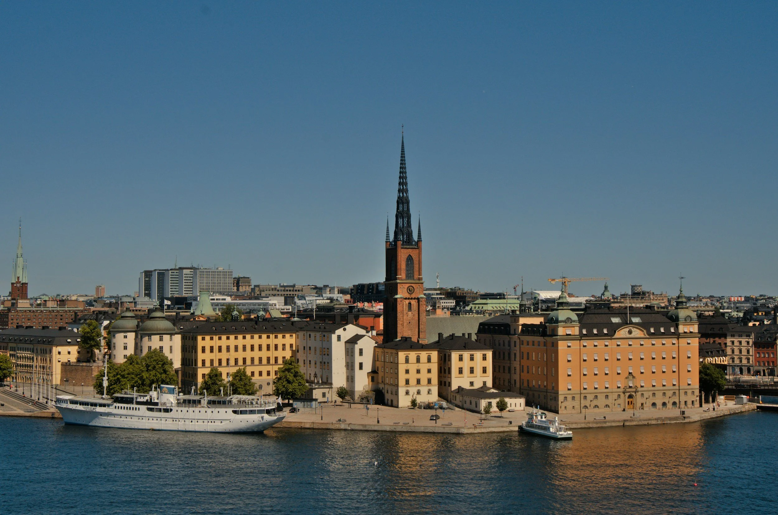 Utsikt över en stad med en kyrktorn och moderna byggnader vid vattnet, med en segelbåt i fordonet och klar himmel.
