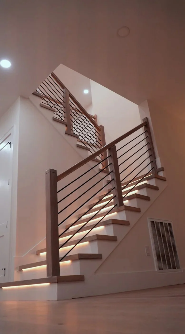 Interior view of a modern staircase with wooden handrails and horizontal metal balusters, illuminated by built-in LED lighting along the steps.